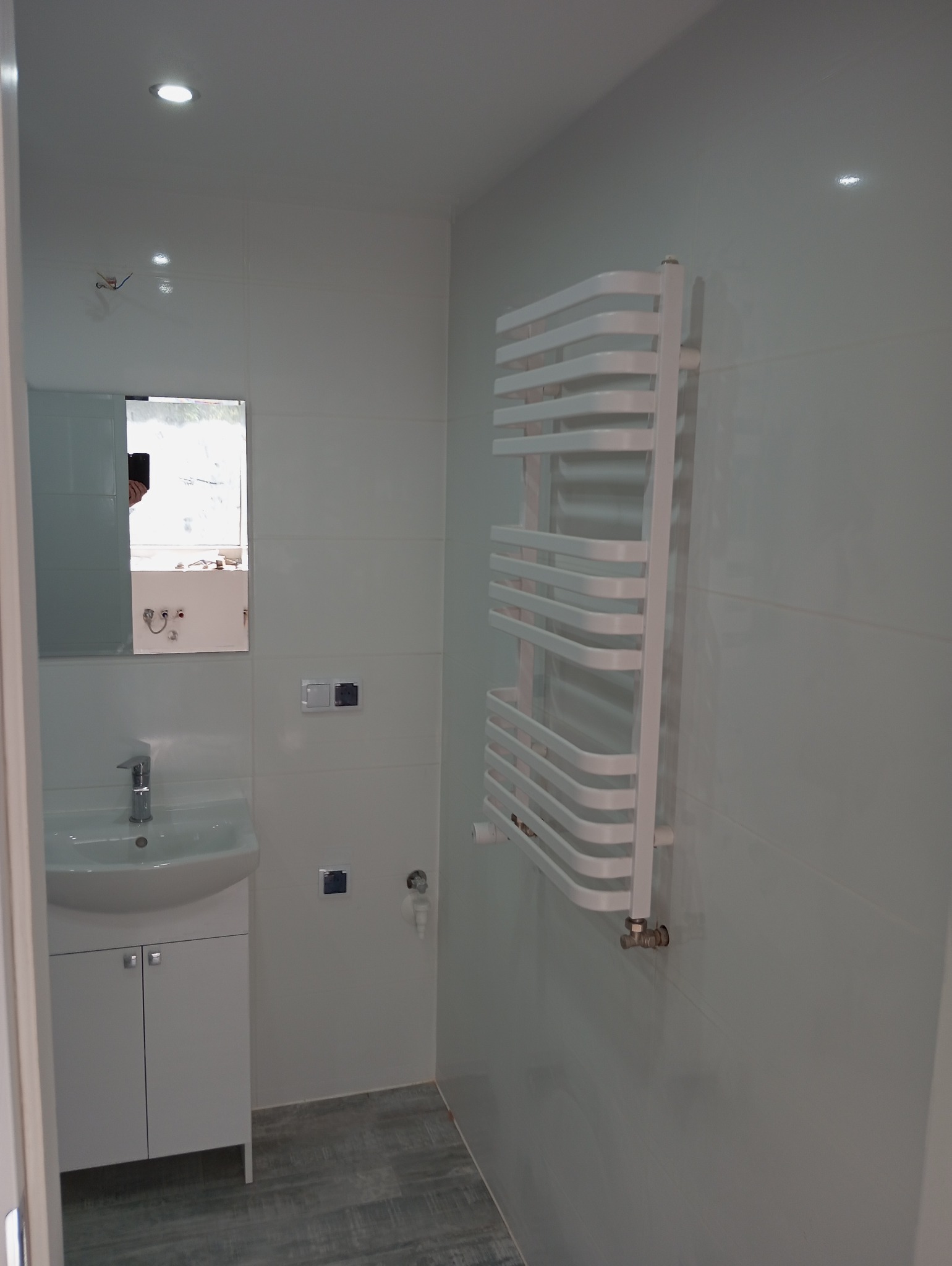 Jasna łazienka po remoncie z umywalką z szafką, grzejnikiem drabinkowym i szarą podłogą imitującą drewno.