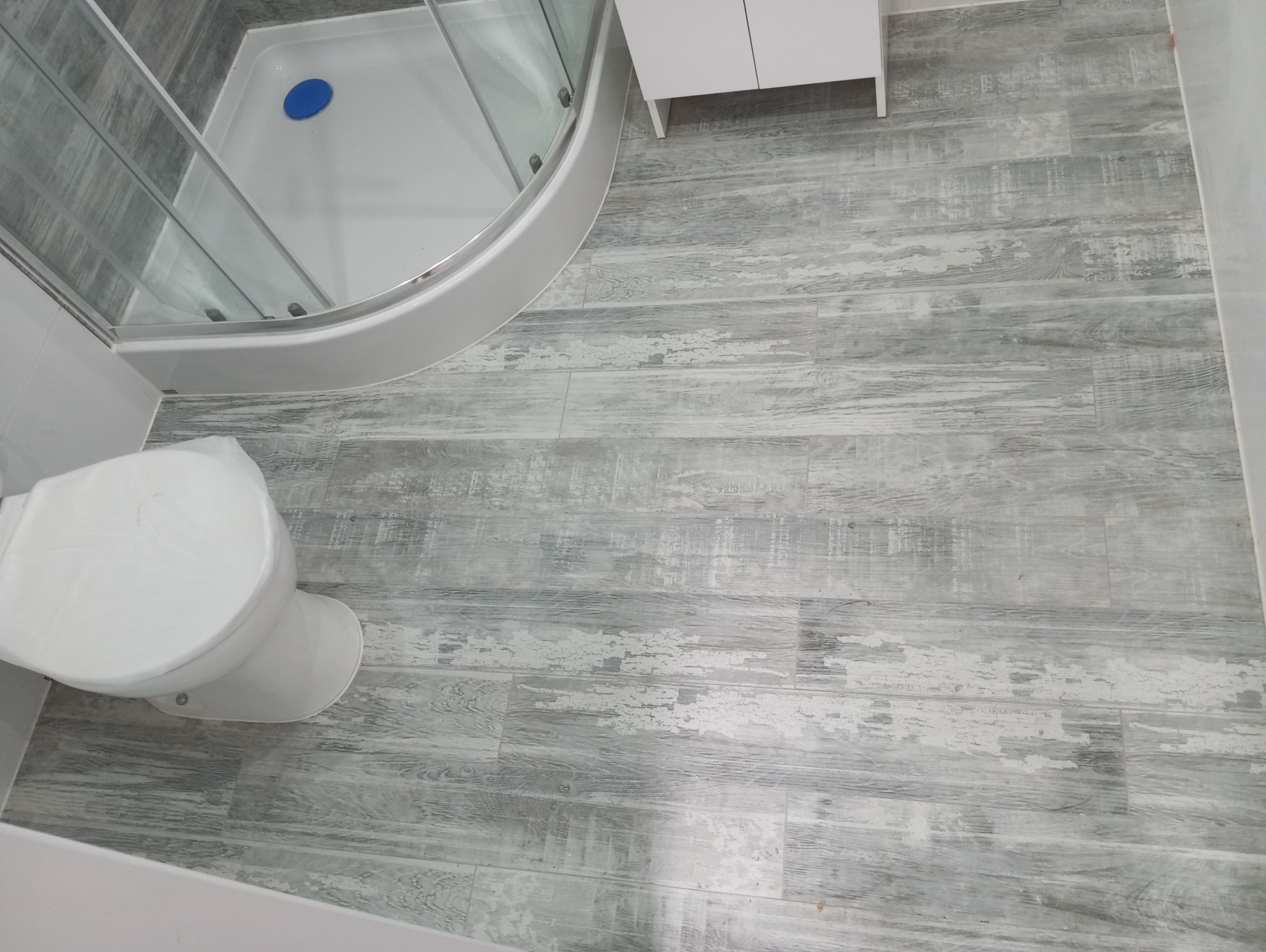 Wykończona łazienka z kabiną prysznicową, białą szafką i toaletą zabezpieczoną folią ochronną, podłoga wyłożona szarymi panelami imitującymi drewno.