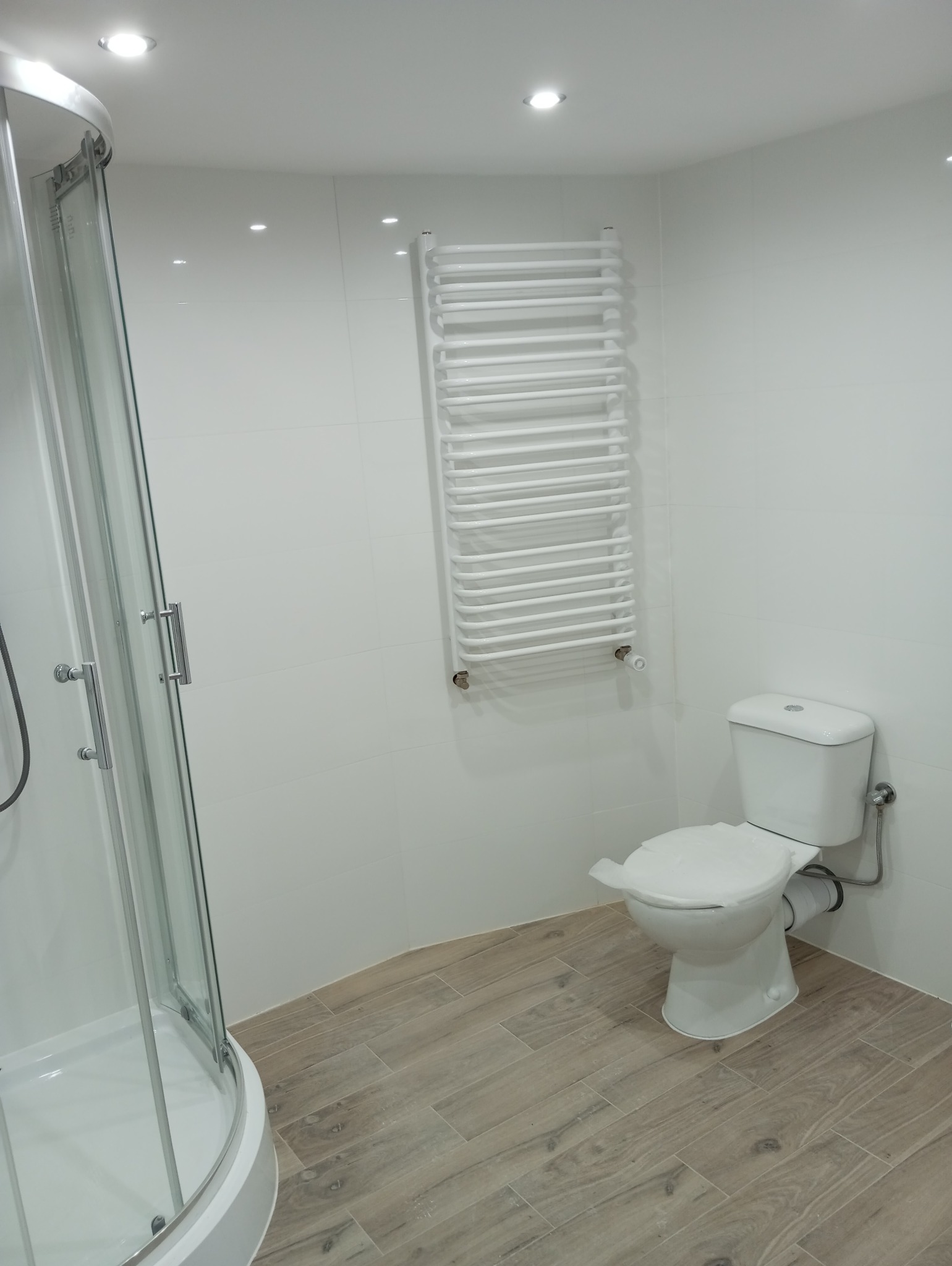 Nowoczesna, jasna łazienka po remoncie z kabiną prysznicową narożną, grzejnikiem drabinkowym i toaletą, wykończona białymi płytkami i podłogą imitującą drewno.