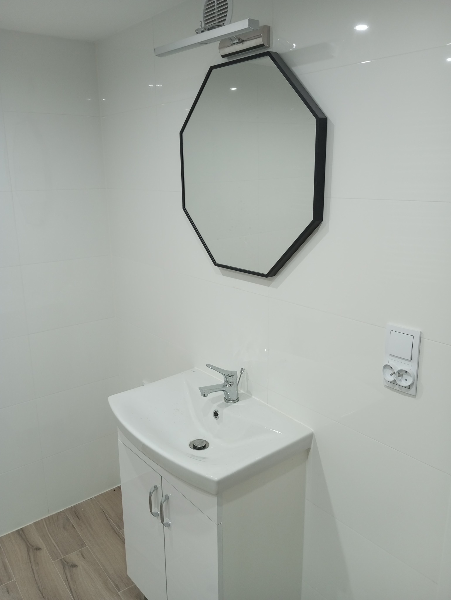 Nowoczesna łazienka z białą umywalką szafkową, ośmiokątnym lustrem w czarnej ramie i oświetleniem punktowym, wykończona białymi płytkami i drewnopodobną podłogą.