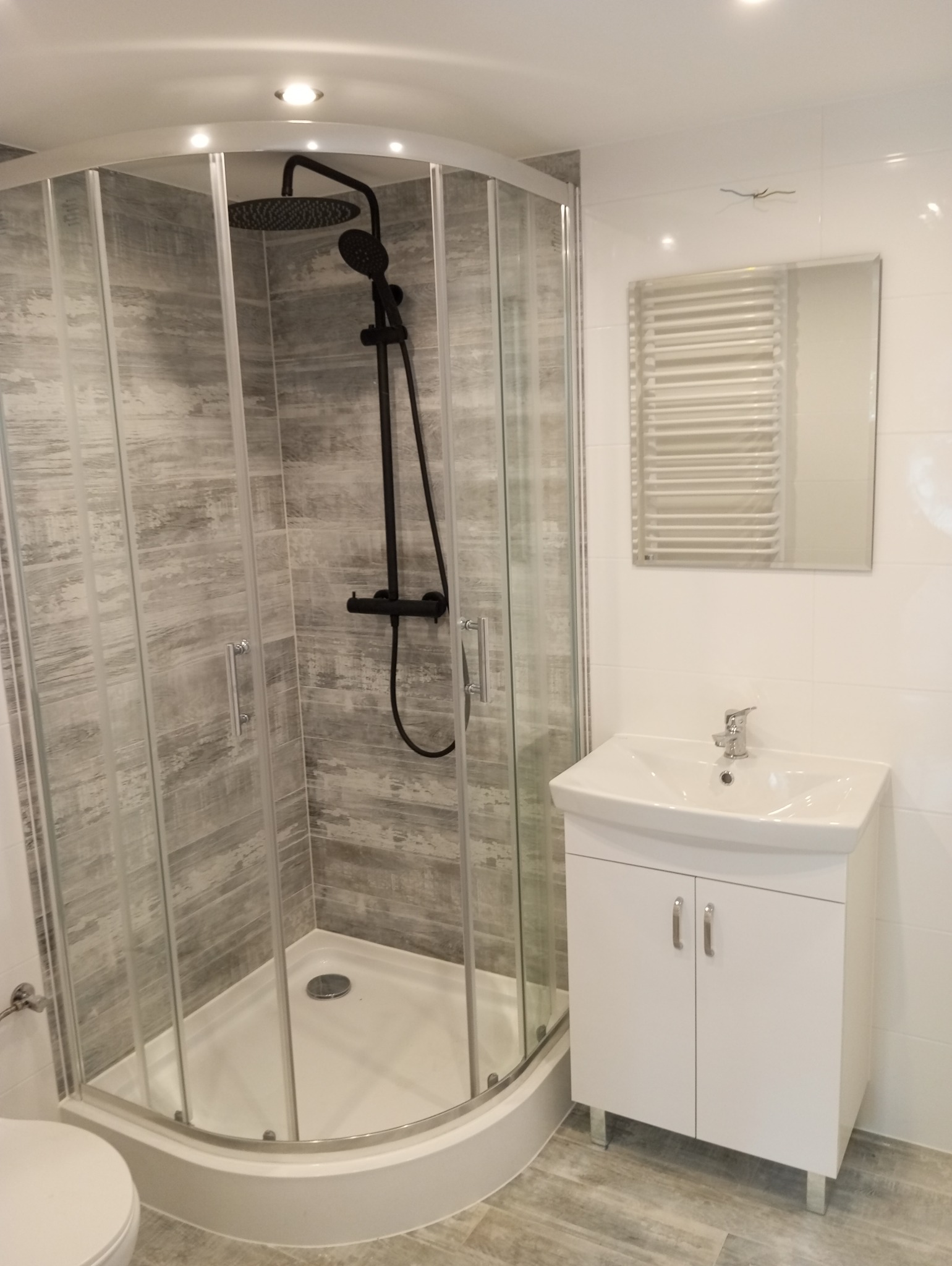 Nowoczesna łazienka z kabiną prysznicową z czarną armaturą, białą umywalką z szafką i grzejnikiem drabinkowym na białych płytkach.