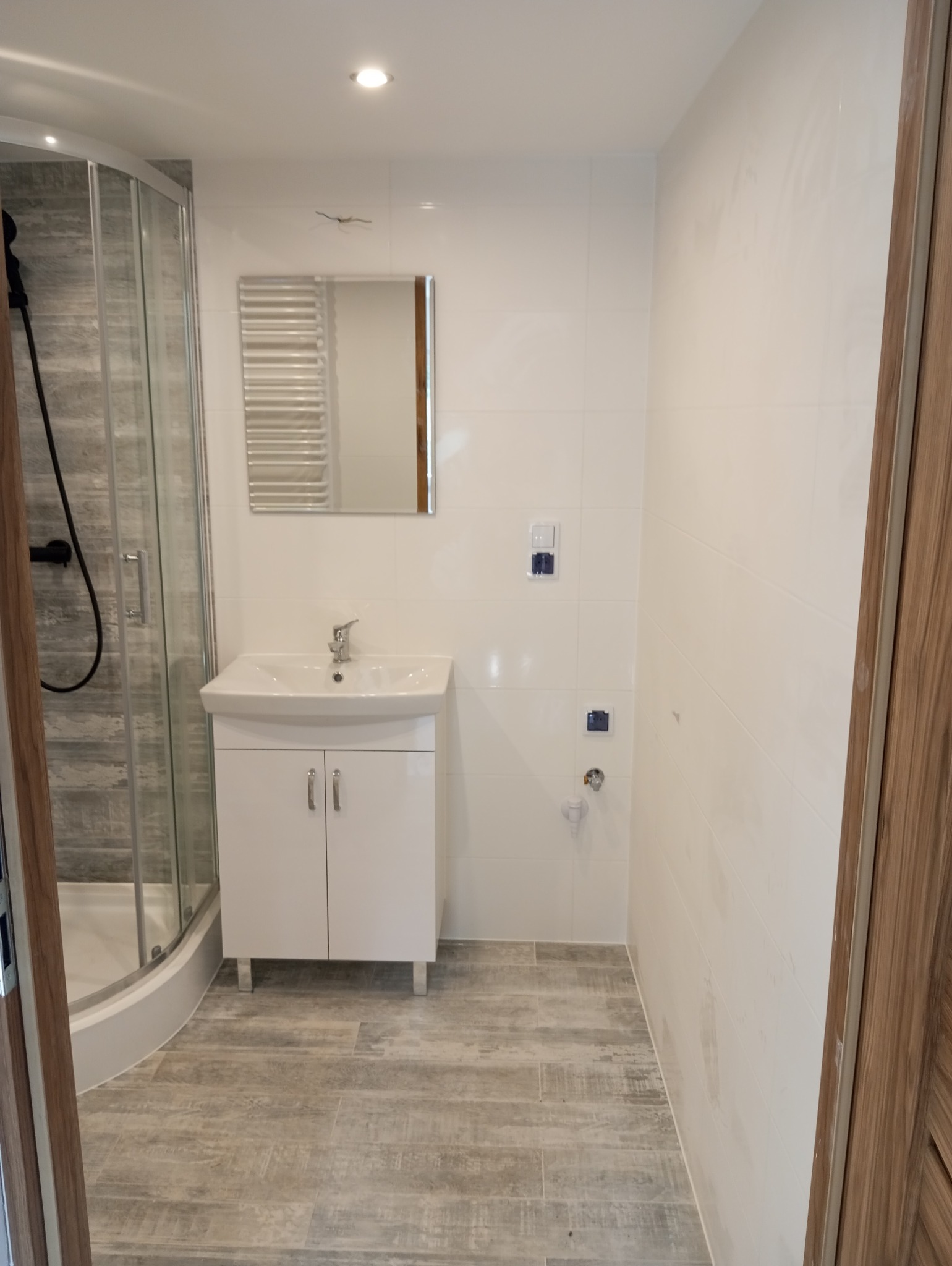 Nowoczesna łazienka w trakcie wykańczania: białe płytki ścienne, kabina prysznicowa z szarymi panelami, biała szafka pod umywalką, lustro i podłoga imitująca drewno, widoczne ślady po pracach...