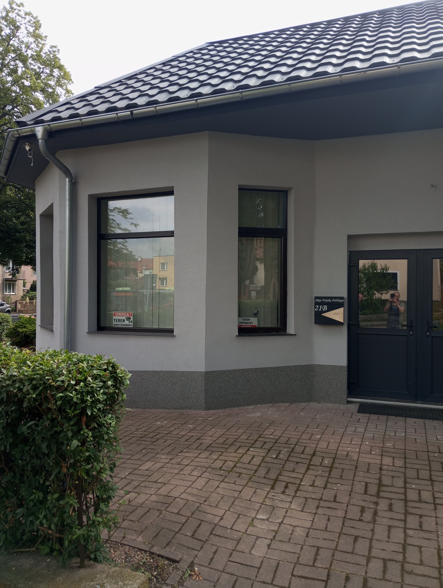 Wykończony budynek z szarą elewacją, ciemną stolarką okienną i nowym dachem z blachodachówki, widoczne tabliczki informacyjne na oknach i skrzynka pocztowa przy drzwiach.