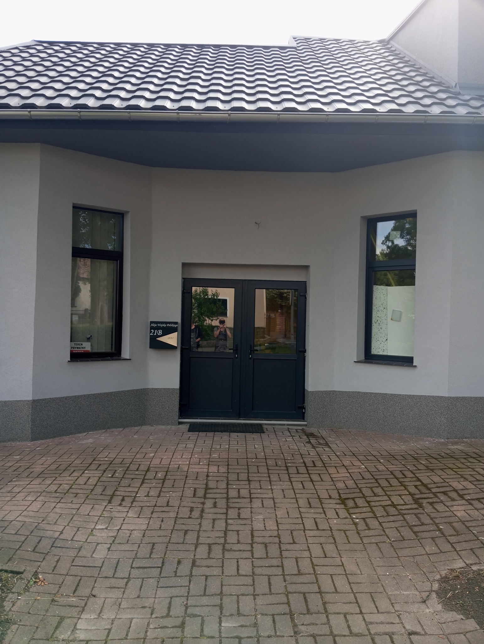Elewacja budynku z szarym tynkiem, ciemnymi oknami i drzwiami wejściowymi, widoczna skrzynka pocztowa z numerem 21B.