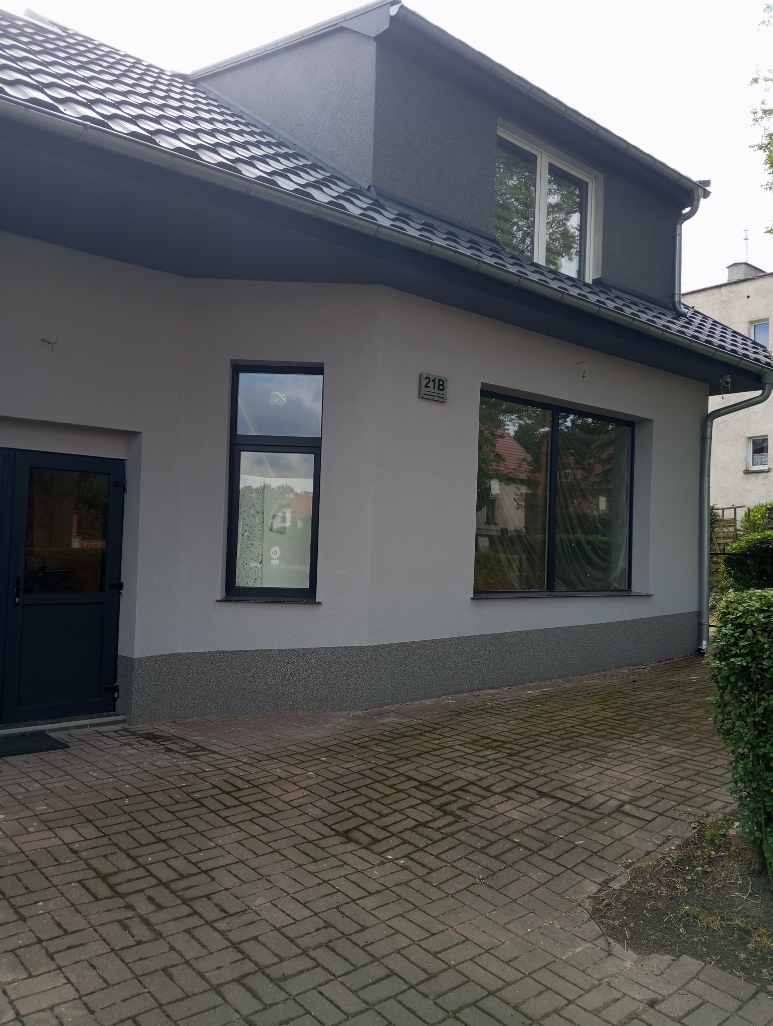 Szary budynek po remoncie elewacji, z ciemnymi oknami i drzwiami, widoczny z zewnątrz na tle kostki brukowej.