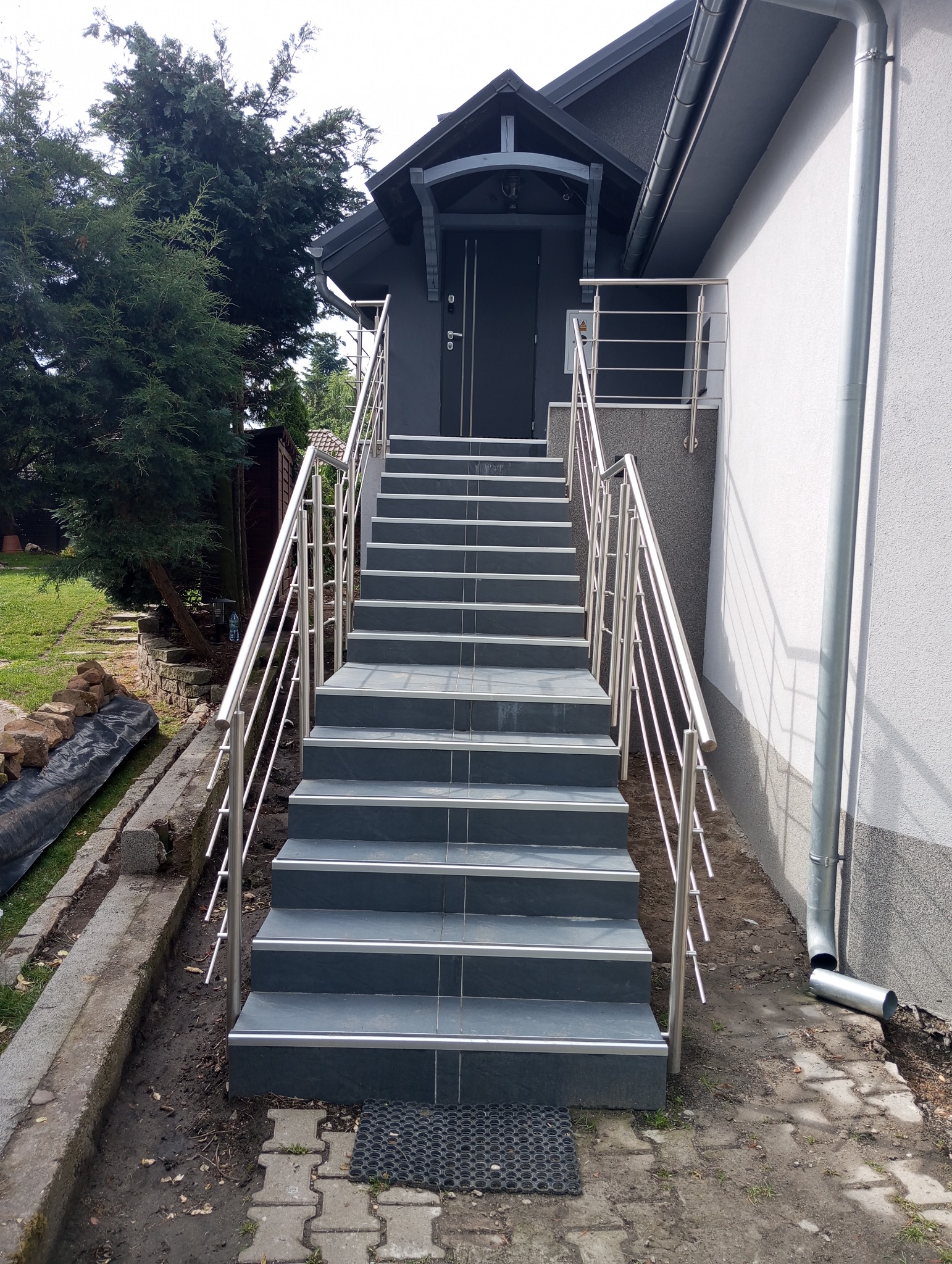 Zewnętrzne schody wejściowe wyłożone grafitową płytką z metalową barierką, prowadzące do ciemnoszarych drzwi wejściowych z zadaszeniem