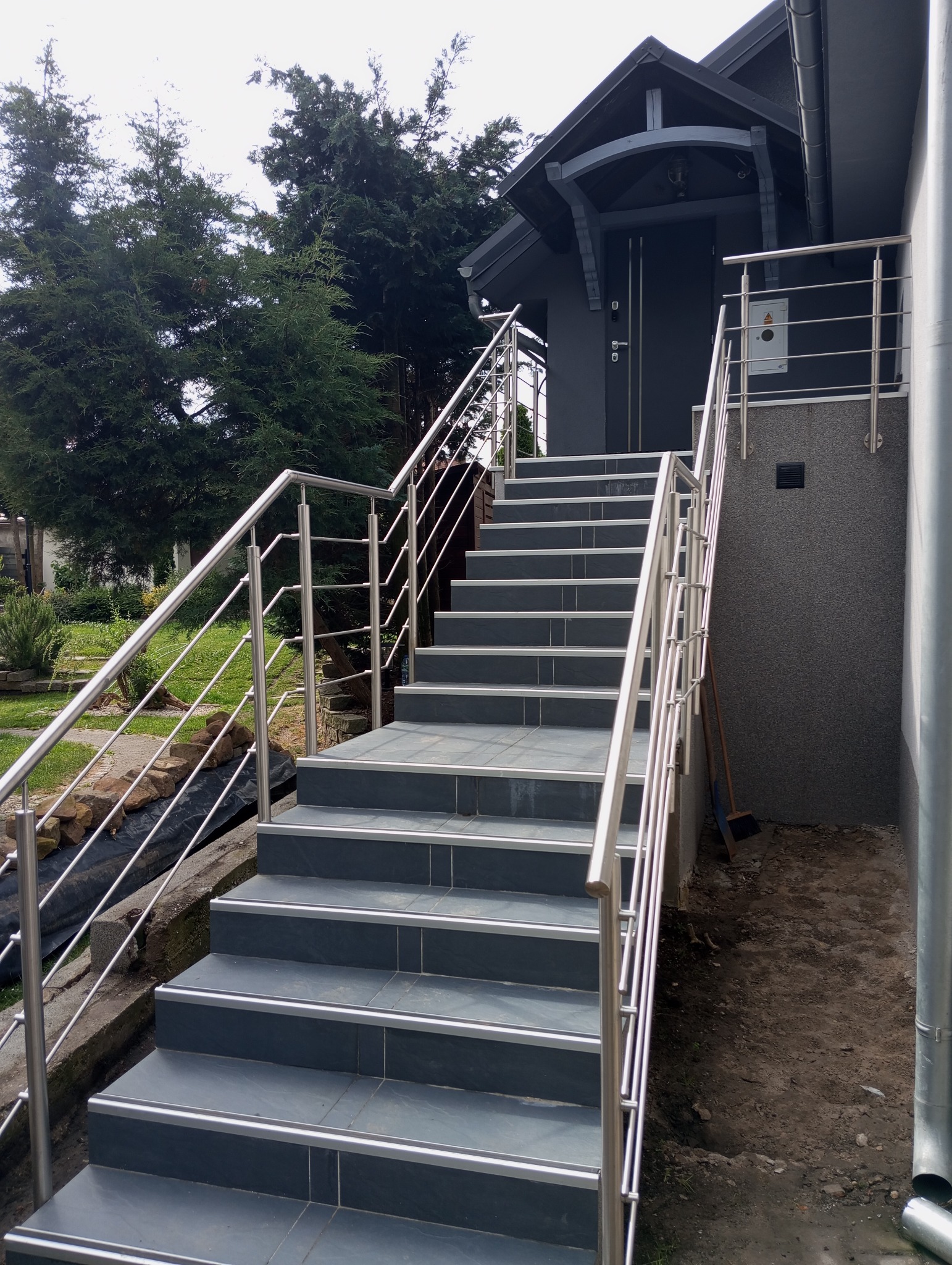 Zewnętrzne schody wejściowe wyłożone szarą płytką z metalowymi krawędziami i balustradą ze stali nierdzewnej prowadzące do szarego budynku z zadaszeniem.