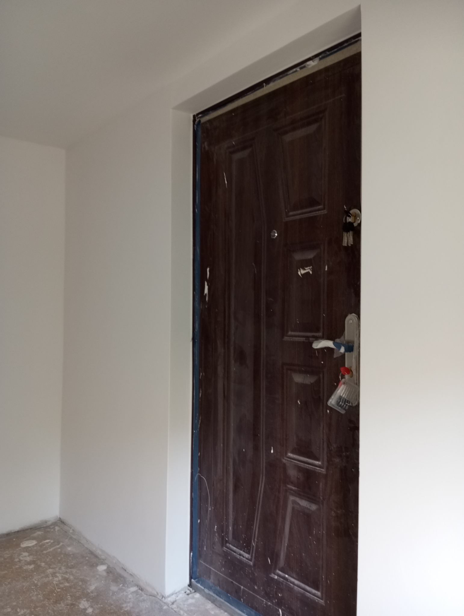Świeżo pomalowane, białe ściany w pomieszczeniu z ciemnymi drzwiami wejściowymi zabezpieczonymi taśmą malarską, widoczne ślady farby na drzwiach.