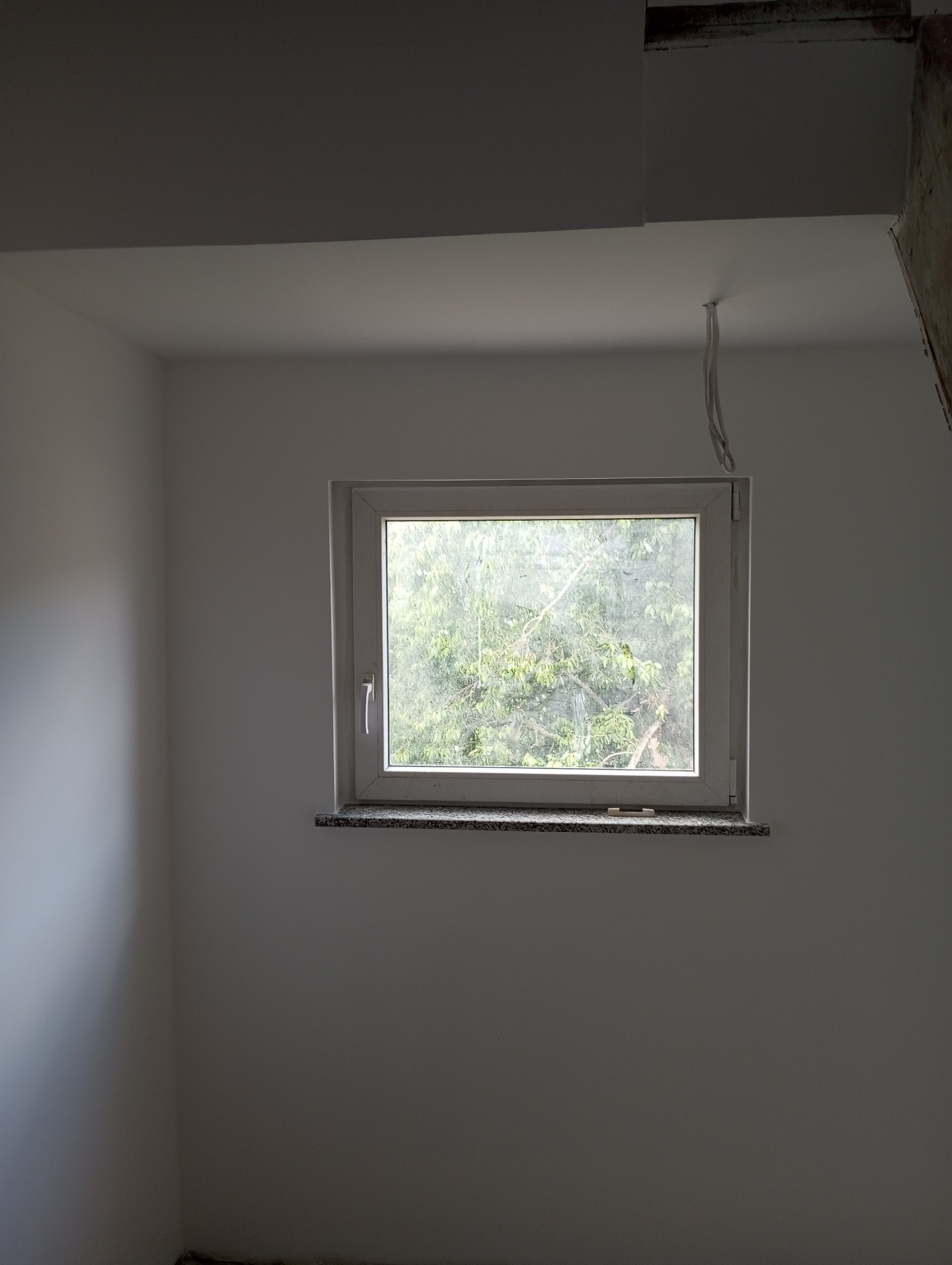 Wnętrze pokoju po malowaniu ścian na biało, widoczne okno z kamiennym parapetem i wystającymi przewodami elektrycznymi z sufitu.