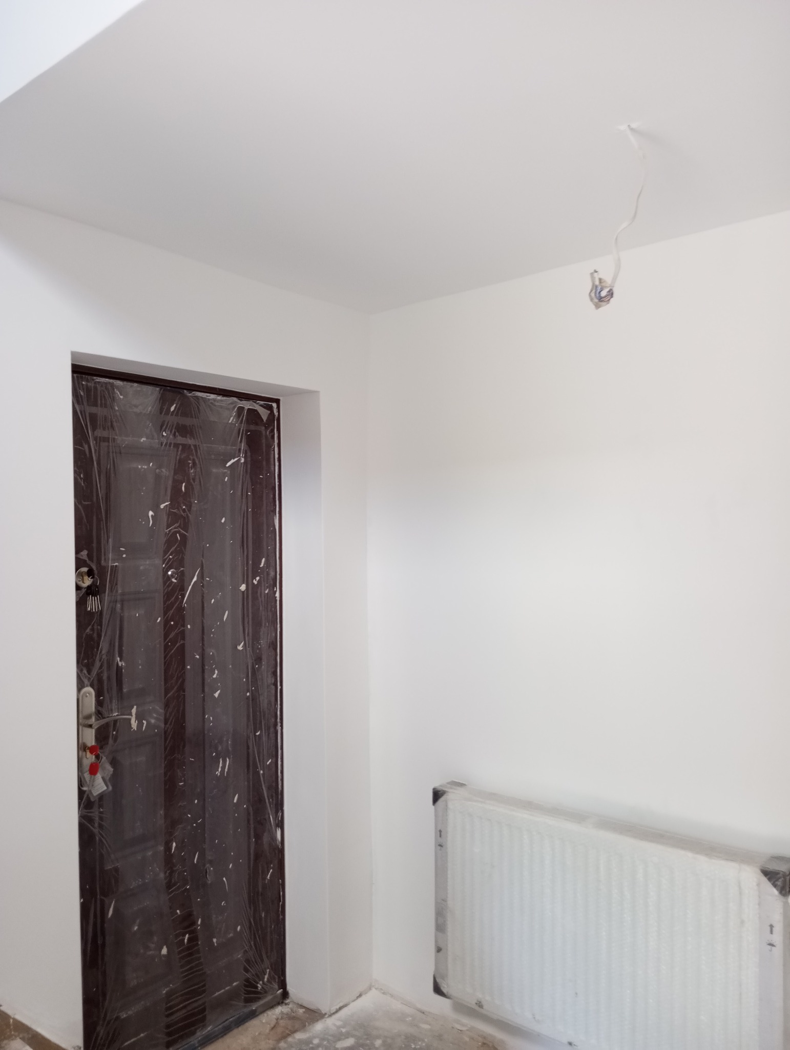 Świeżo pomalowane na biało wnętrze z zabezpieczoną folią brązową ościeżnicą, grzejnikiem i wystającymi przewodami elektrycznymi.