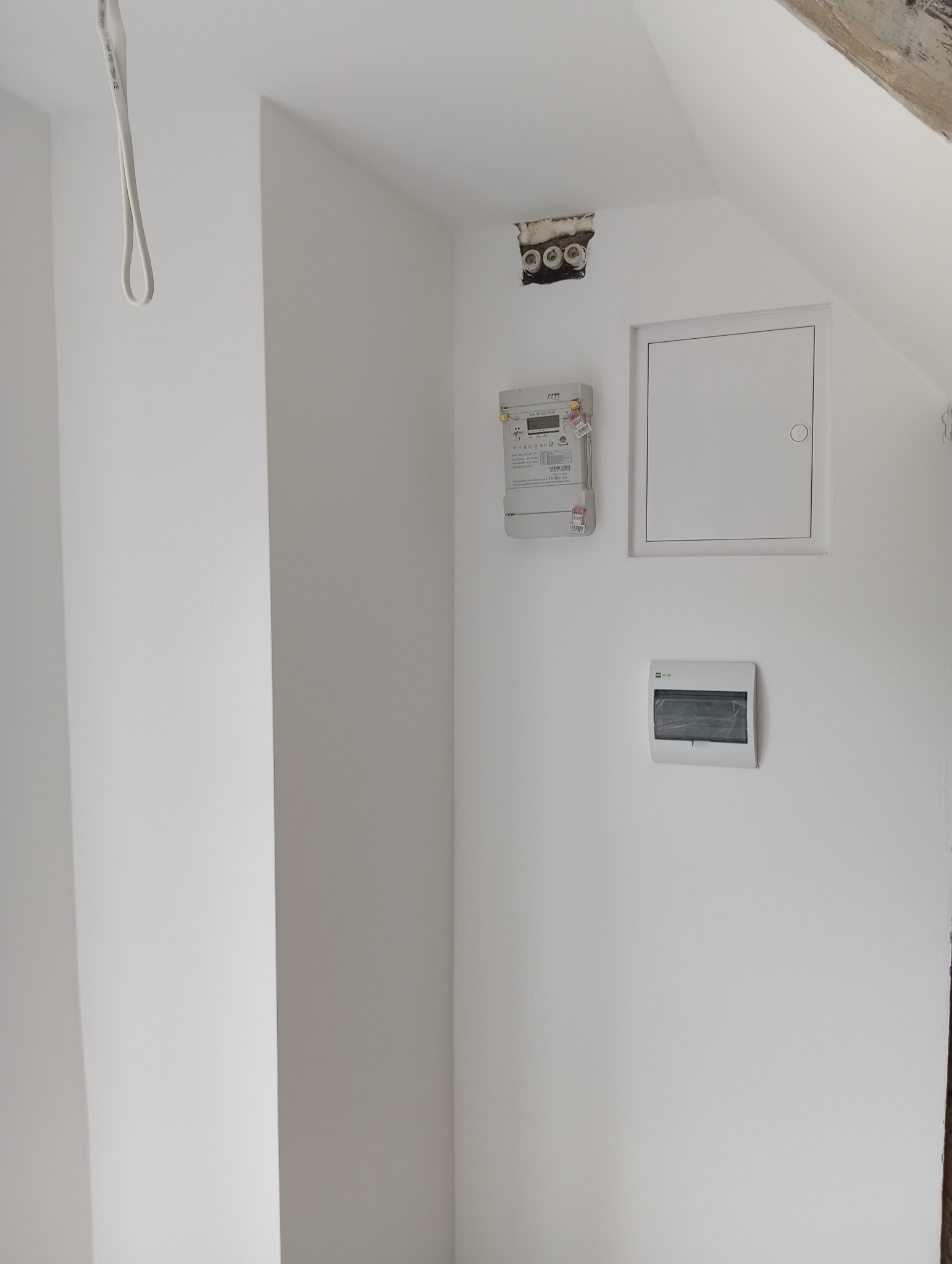 Świeżo pomalowana na biało ściana poddasza z widocznymi elementami instalacji elektrycznej: otwarta puszka z przewodami, licznik energii, skrzynka elektryczna i klapa rewizyjna.