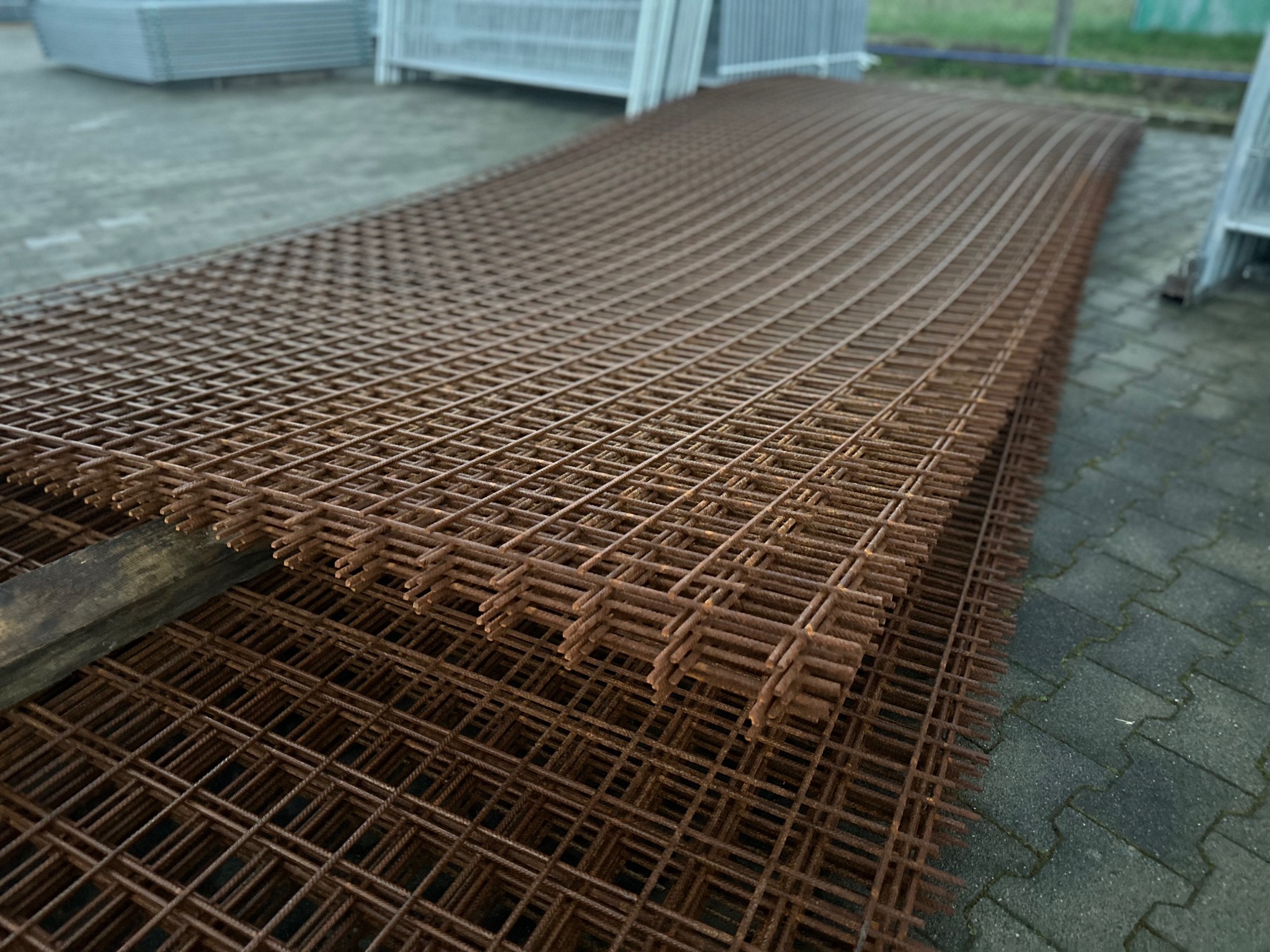 Stos brązowych, zardzewiałych paneli ogrodzeniowych z metalowej siatki, ułożonych na drewnianej belce, na tle betonowej kostki brukowej i innych paneli ogrodzeniowych.