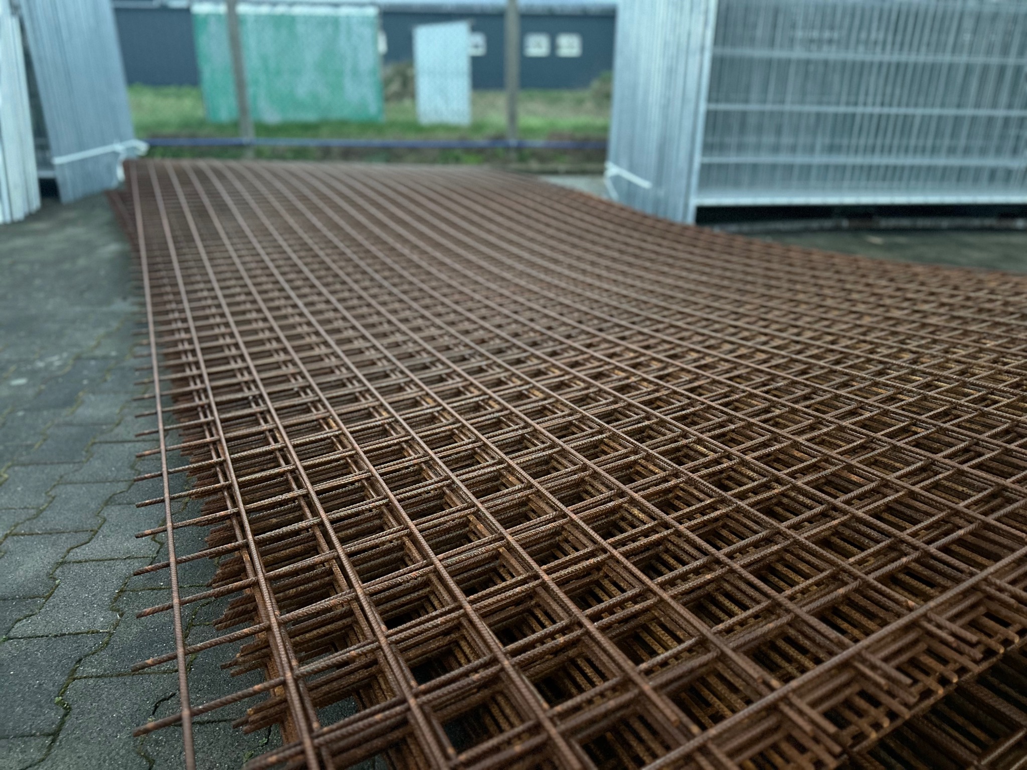 Stos zardzewiałych, kwadratowych paneli siatkowych na chodniku z kostki brukowej, w tle częściowo widoczne elementy ogrodzenia panelowego.