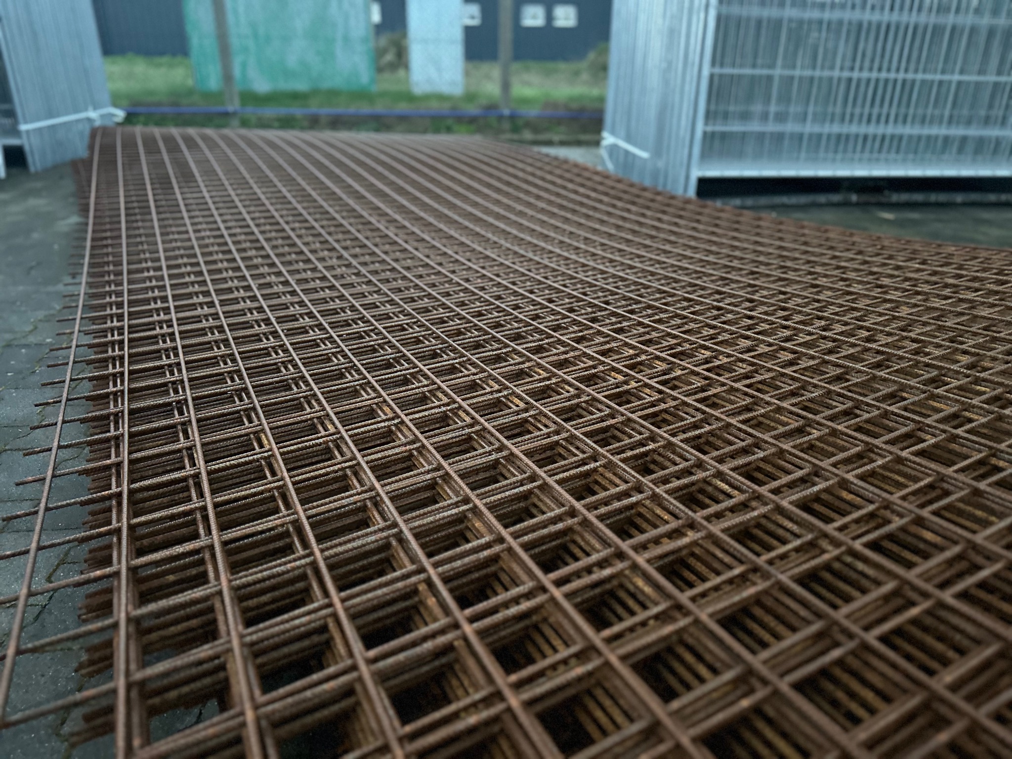 Stos brązowych, zardzewiałych paneli ogrodzeniowych zgrzewanych punktowo, ułożonych warstwami na betonowej posadzce, z widocznymi elementami ocynkowanych przęseł w tle.