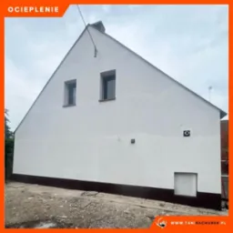 Dom jednorodzinny z białymi ścianami po remoncie, widoczne dwa okna na poddaszu i brązowa podmurówka, teren wokół domu w trakcie prac wykończeniowych.