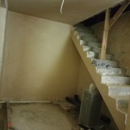 MAXBUD MARIUSZ ROJEK - Wnętrze po nałożeniu tynków: gładkie ściany w odcieniach beżu, surowe schody betonowe z drewnianą podporą, widoczne przewody i materiały budowlane na podłodze.