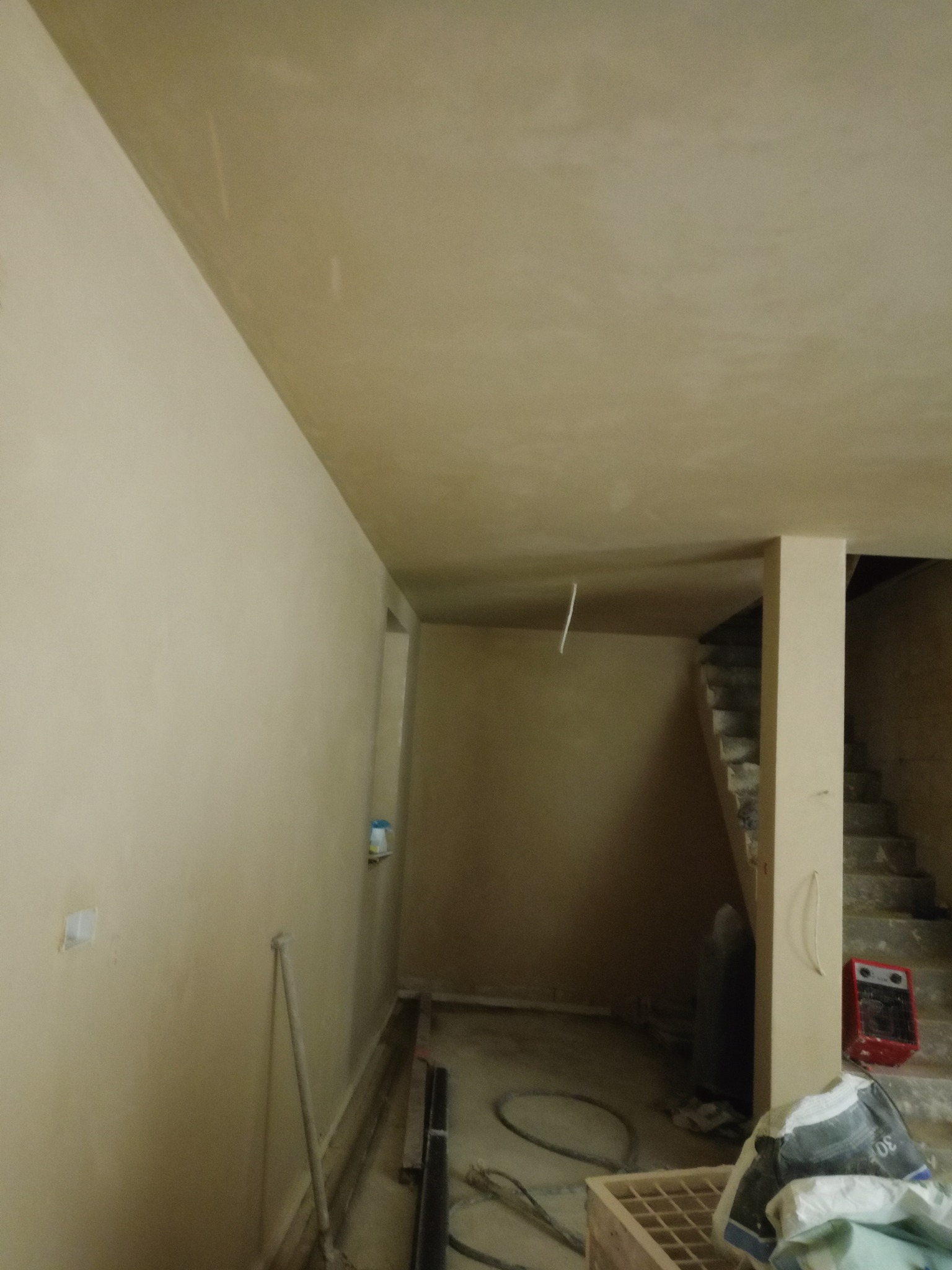 Wnętrze po nałożeniu tynków: gładkie, beżowe ściany i sufit, widoczne schody w tle, narzędzia i materiały budowlane na podłodze. Minimalistyczny efekt po pracach wykończeniowych.