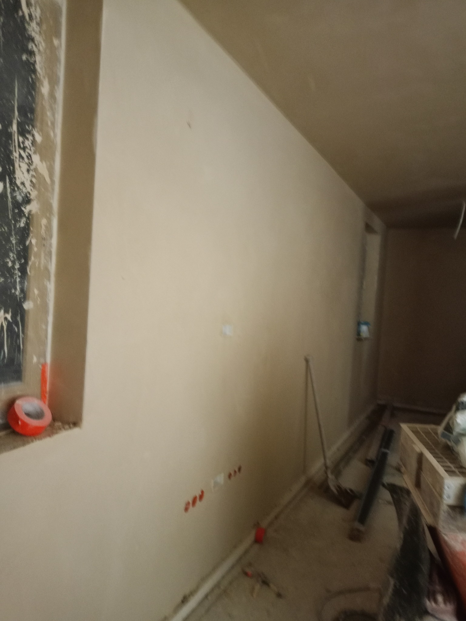 Gładka, beżowa ściana po nałożeniu tynku maszynowego, widoczne ślady po gniazdkach elektrycznych i taśma malarska przy oknie. W tle narzędzia i materiały budowlane.