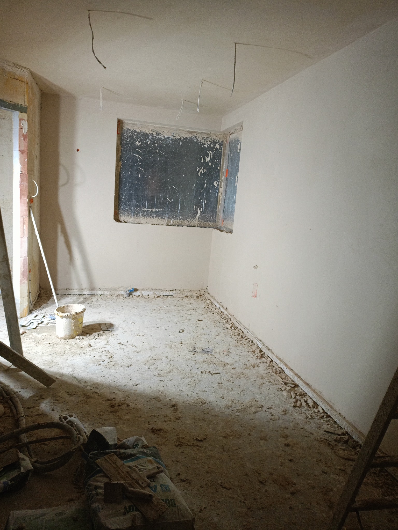 Pokój po tynkowaniu maszynowym: gładkie, jasne ściany, resztki zaprawy na podłodze, widoczne kable i drabina. Okno zabezpieczone folią.