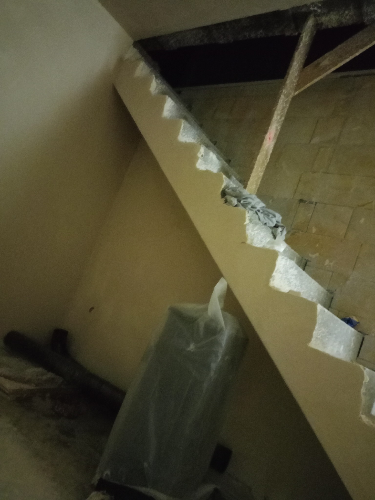 Niedokończone tynkowanie ściany pod skosem, widoczne fragmenty starego muru i elementy konstrukcji dachu. Na podłodze rury i folia ochronna.