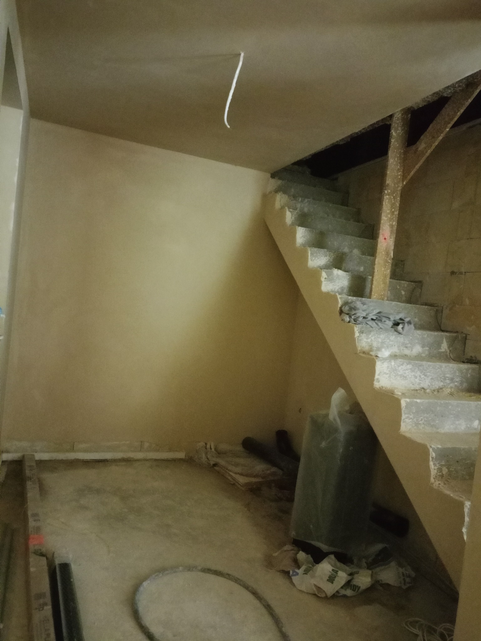 Wnętrze po nałożeniu tynków: gładkie ściany w odcieniach beżu, surowe schody betonowe z drewnianą podporą, widoczne przewody i materiały budowlane na podłodze.