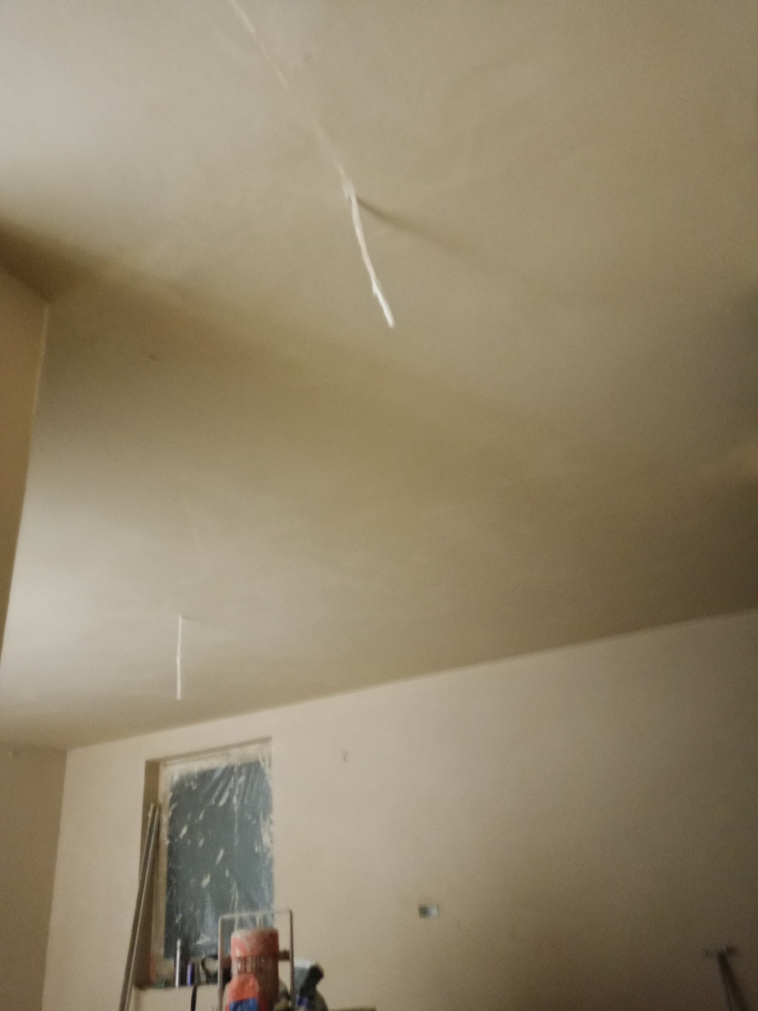 Gładka, jasna powierzchnia świeżo nałożonego tynku na ścianach i suficie w pokoju. Okno zabezpieczone folią. Widoczny sprzęt do tynkowania.