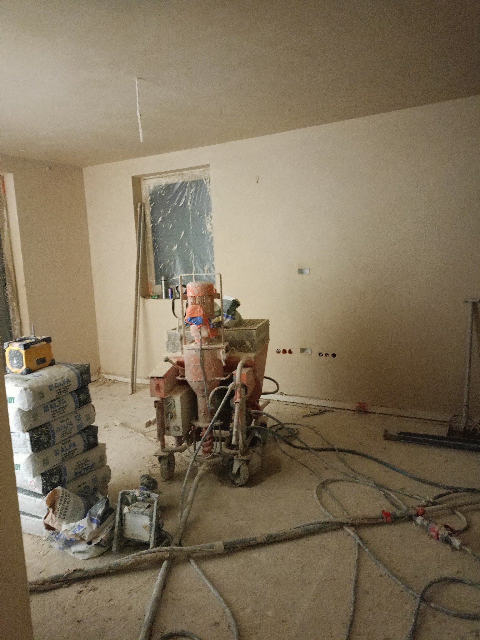 Wnętrze po nałożeniu tynków maszynowych. Widoczna maszyna do tynkowania, worki z zaprawą i świeżo otynkowane ściany w jasnym odcieniu beżu. Okno zabezpieczone folią.