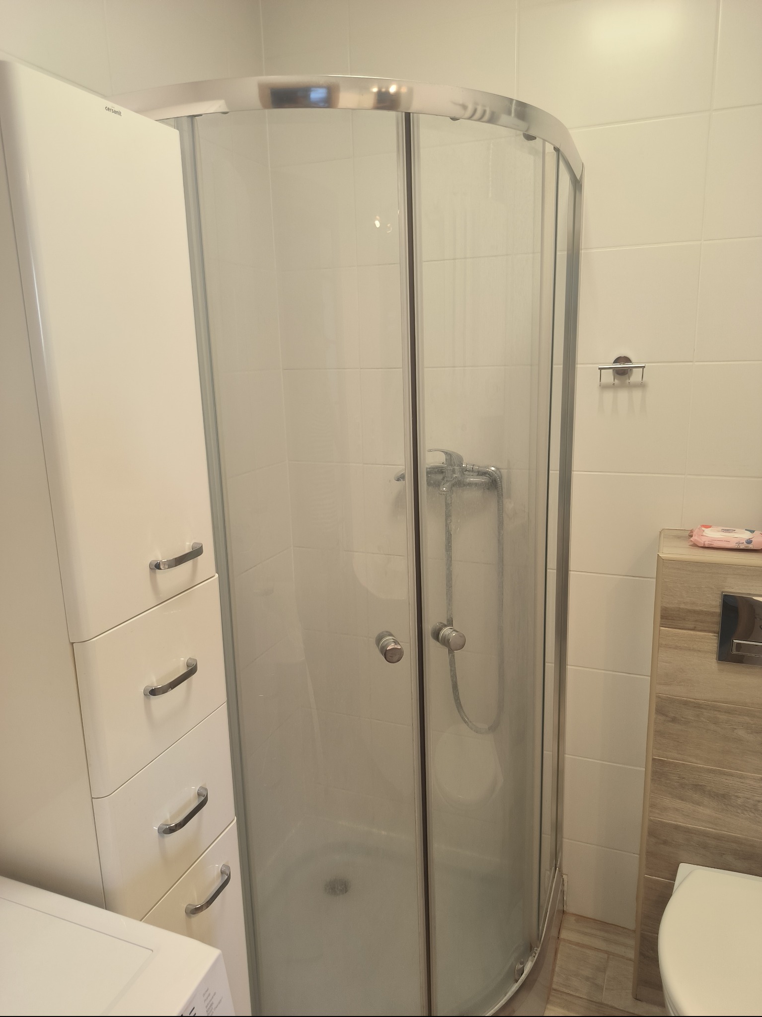 Błyszcząca kabina prysznicowa z chromowanymi elementami w łazience z białymi płytkami, widoczny biały słupek z szufladami i fragment pralki.