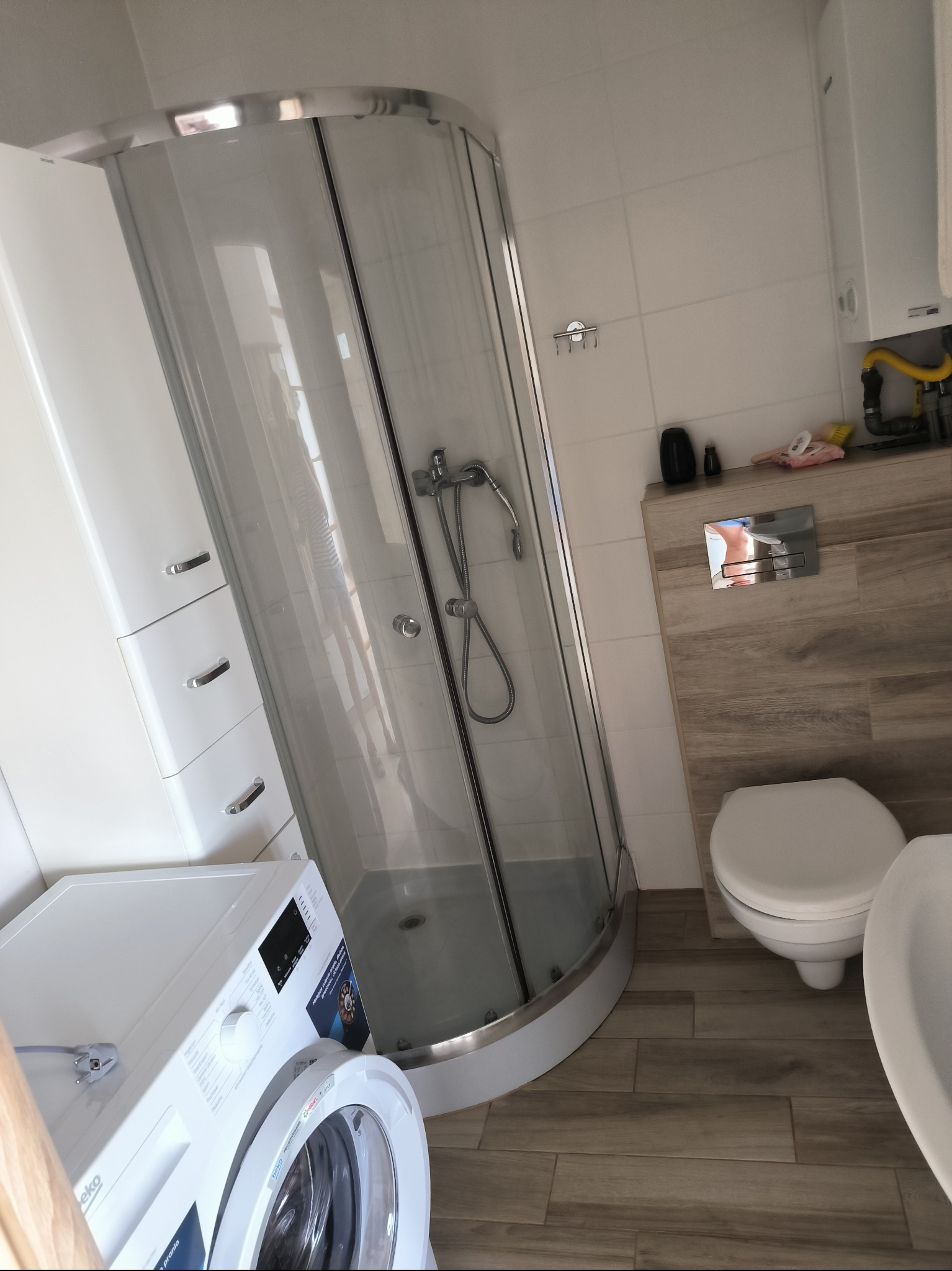 Biała łazienka z kabiną prysznicową, pralką Beko oraz toaletą, wykończona płytkami imitującymi drewno.