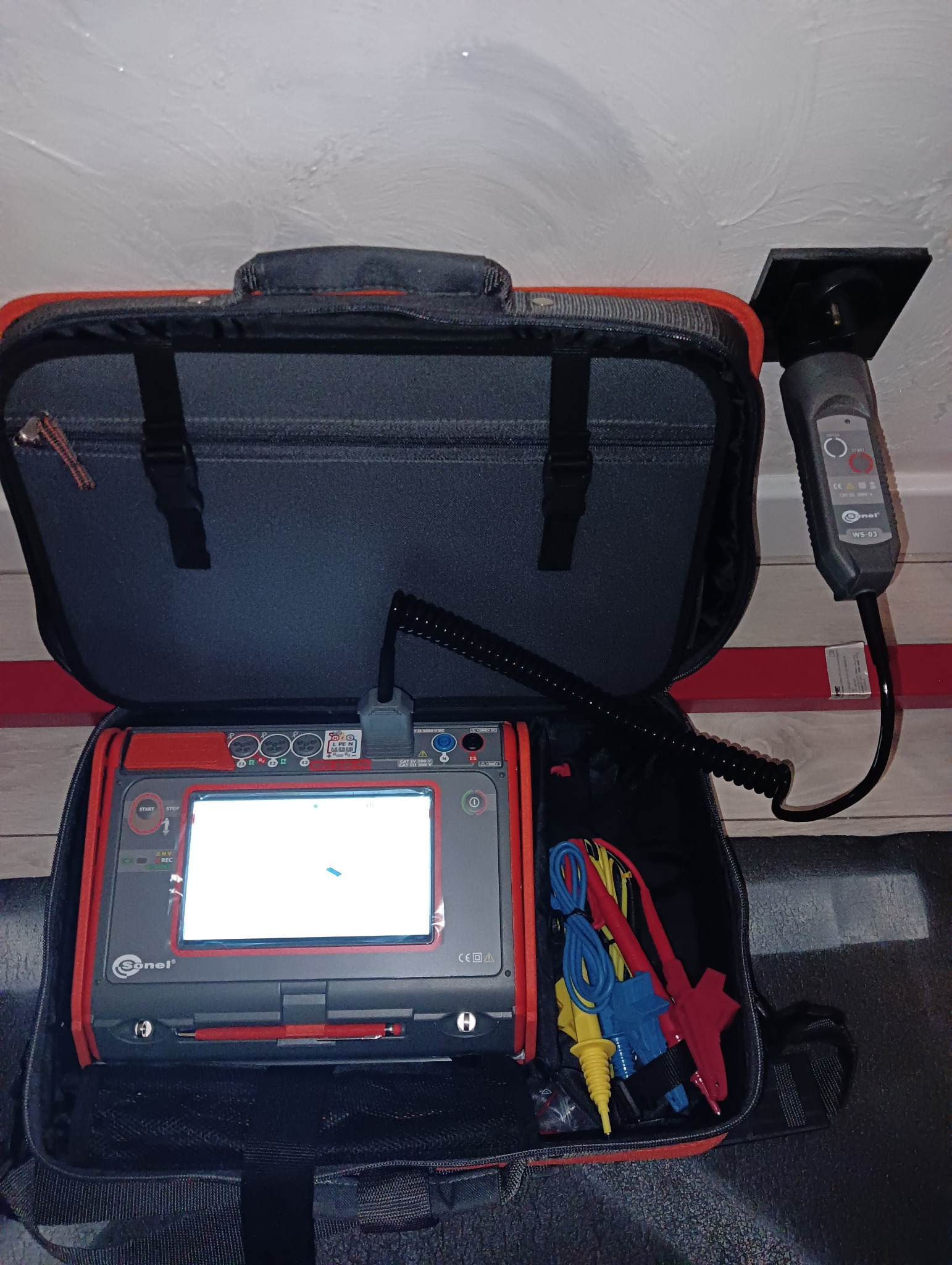 Otwarta torba transportowa z miernikiem elektrycznym Sonel, widoczne przewody pomiarowe, dodatkowy czujnik przymocowany do sufitu.