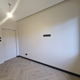 Maciej Smreczak - Wykończone wnętrze pokoju z podłogą ułożoną w jodełkę, świeżo pomalowanymi ścianami w odcieniach bieli i zamontowanymi czarnymi listwami przypodłogowymi oraz gniazdkami elektrycznymi.