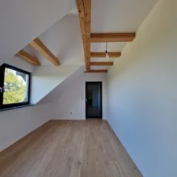 Poddasze po remoncie z drewnianymi belkami stropowymi, świeżo pomalowanymi białymi ścianami, panelami podłogowymi w odcieniach drewna i czarnym oknem z widokiem na zieleń.