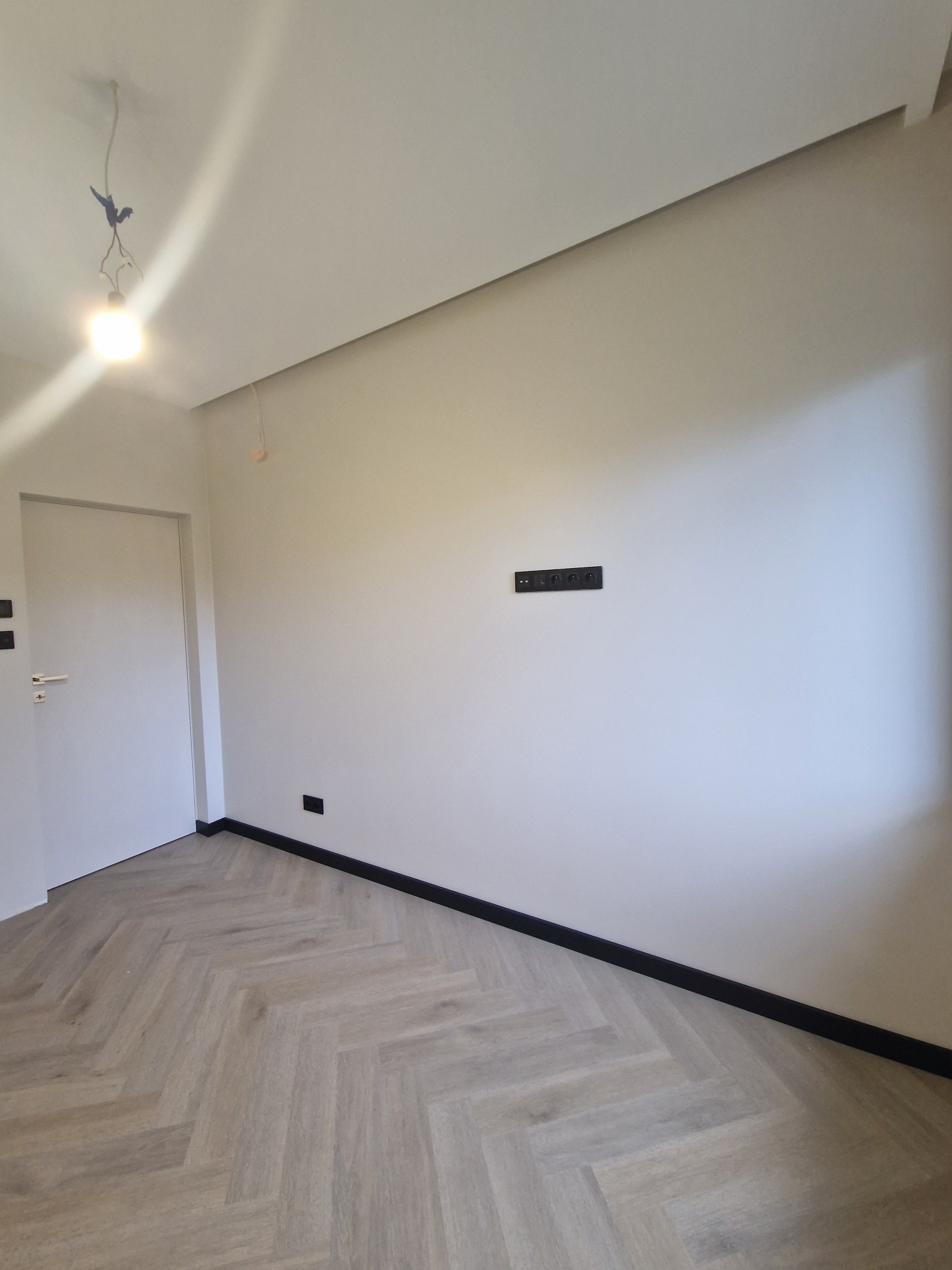 Wykończone wnętrze pokoju z podłogą ułożoną w jodełkę, świeżo pomalowanymi ścianami w odcieniach bieli i zamontowanymi czarnymi listwami przypodłogowymi oraz gniazdkami elektrycznymi.