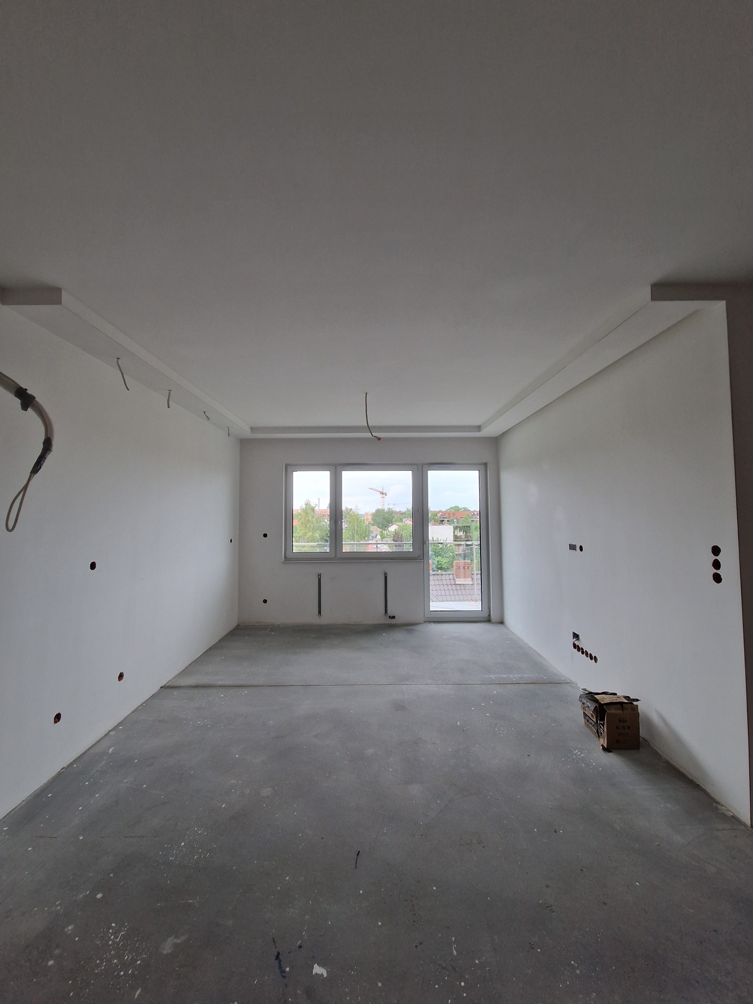 Pusty pokój w trakcie remontu, świeżo pomalowane ściany na biało, surowa betonowa podłoga z resztkami farby, widoczne puszki elektryczne i wystające kable, okno z widokiem na zabudowę jednorodzinną...