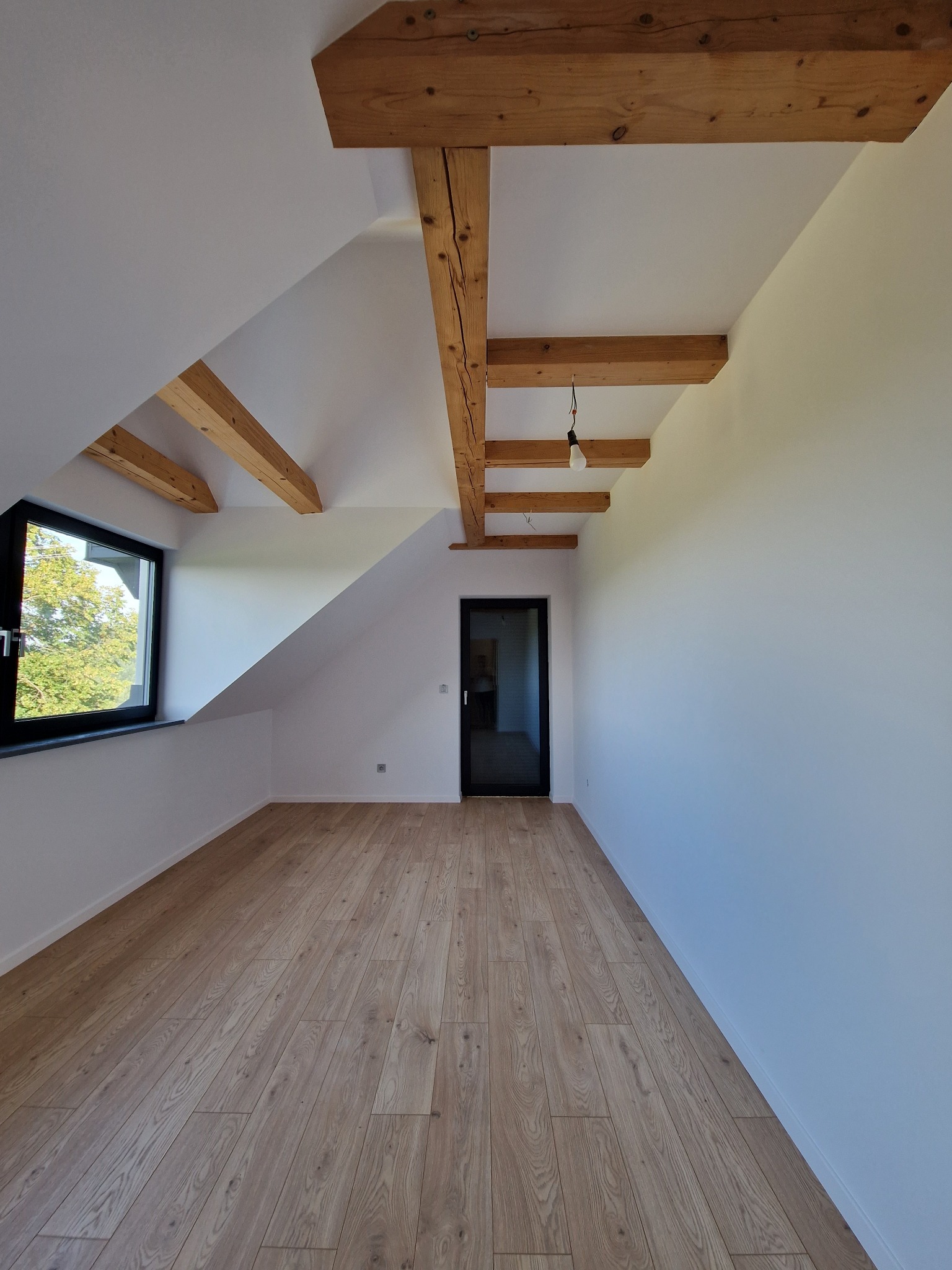 Poddasze po remoncie z drewnianymi belkami stropowymi, świeżo pomalowanymi białymi ścianami, panelami podłogowymi w odcieniach drewna i czarnym oknem z widokiem na zieleń.