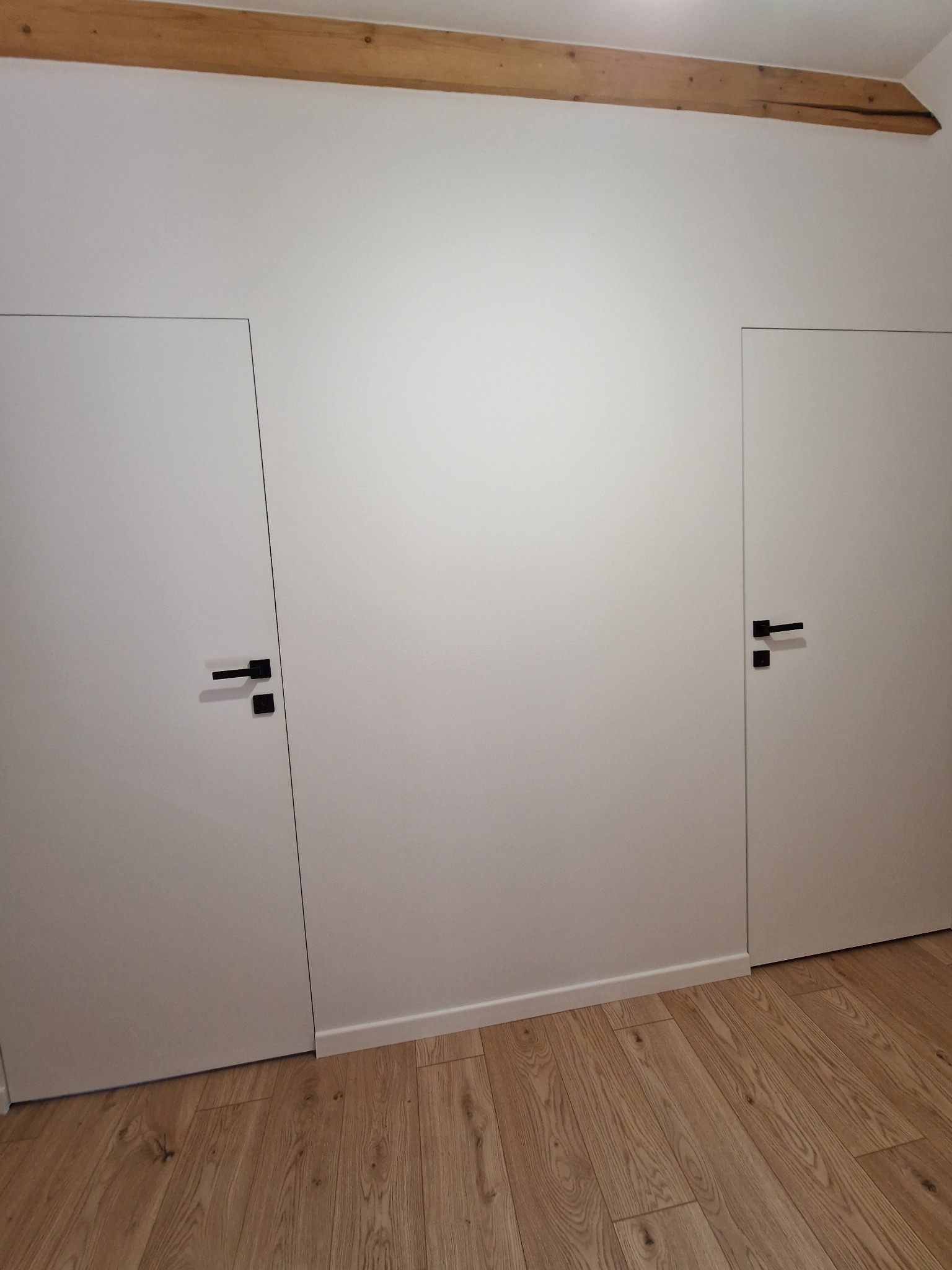 Białe, ukryte drzwi z czarnymi klamkami w białej ścianie poddasza z drewnianą belką stropową, wykończone jasnymi panelami podłogowymi.