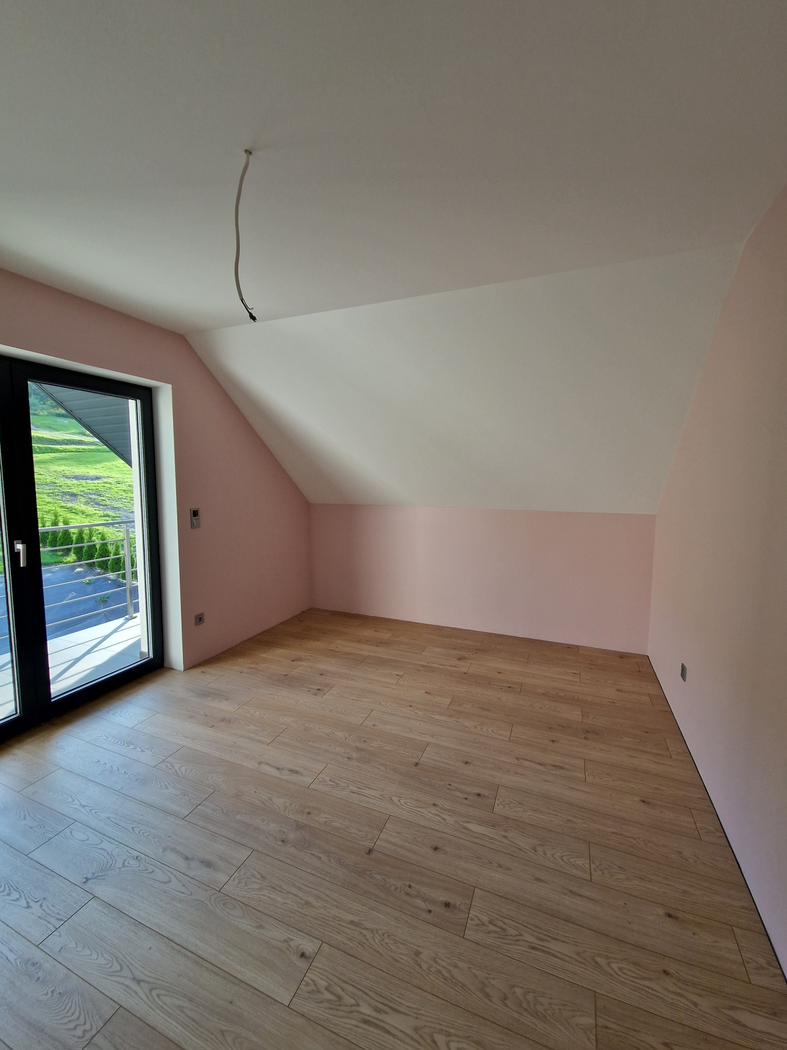 Poddasze po remoncie: ściany pomalowane na różowo, drewniana podłoga, widok na zewnątrz przez czarne drzwi balkonowe, wystający kabel ze sufitu.