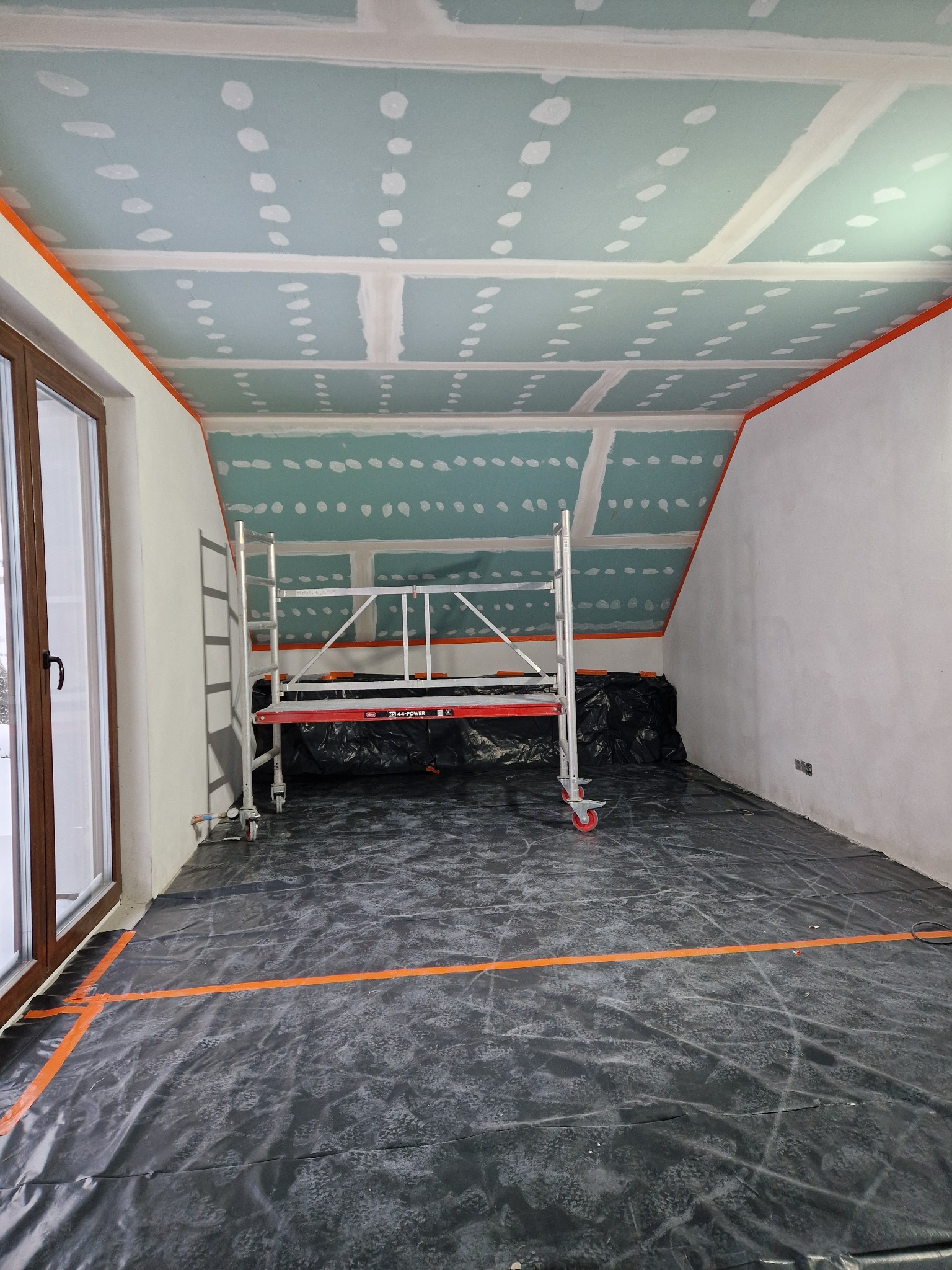 Poddasze w trakcie montażu płyt kartonowo-gipsowych, widoczne rusztowanie, zabezpieczona folią podłoga i taśma malarska na ścianach.