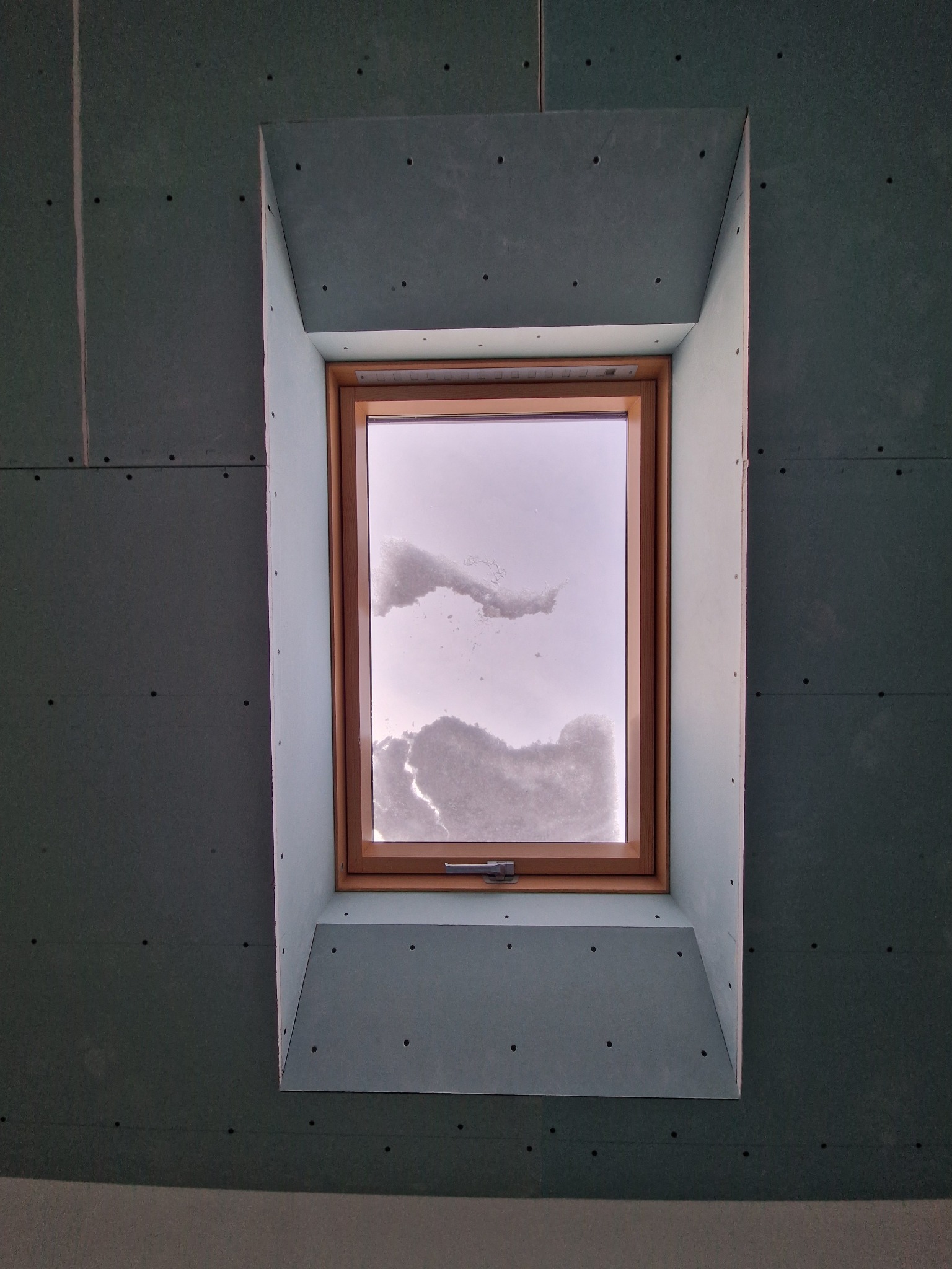 Okno dachowe w trakcie montażu, obudowane płytami gipsowo-kartonowymi, widoczne ślady po wkrętach, za oknem zimowy krajobraz.