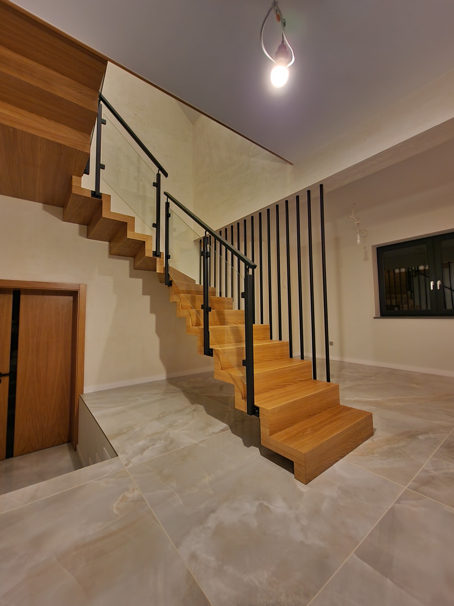 Drewniane schody z czarną balustradą i szklanymi panelami w jasnym wnętrzu z marmurową podłogą i drewnianymi drzwiami, oświetlone wiszącą żarówką.