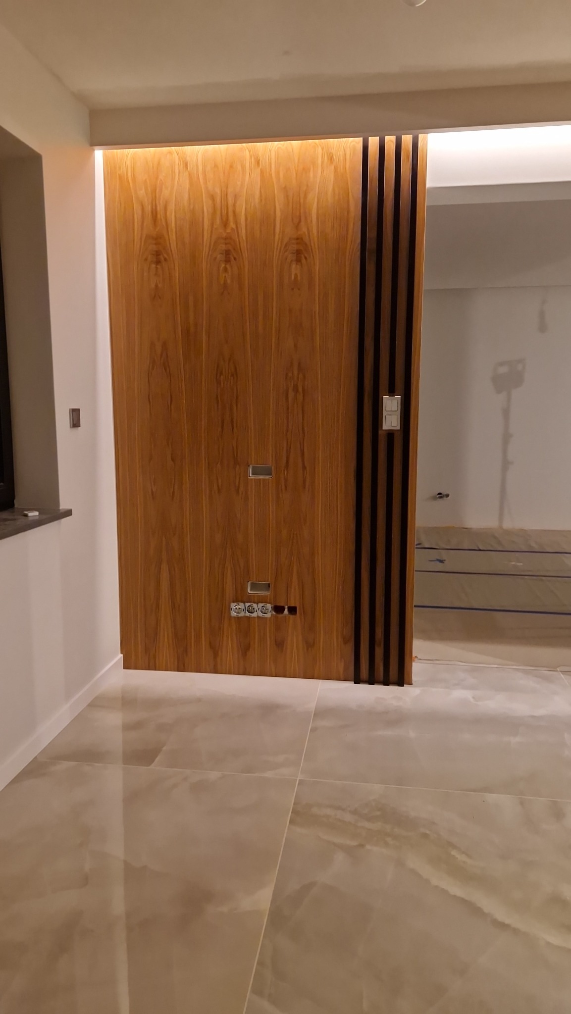 Drewniane drzwi przesuwne z czarnymi pionowymi paskami, widoczne gniazdka elektryczne i włącznik, jasna podłoga z płytek imitujących marmur.