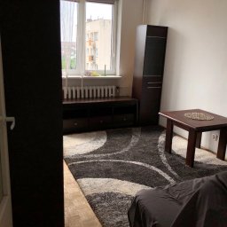 Twój Konsjerż - Umeblowany pokój dzienny z wzorzystym dywanem, drewnianym stolikiem kawowym i szafką RTV pod oknem, widok zza uchylonych drzwi.