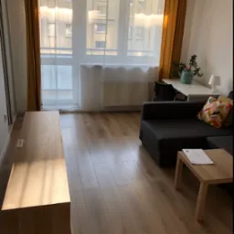 Jasne wnętrze pokoju dziennego z drewnianą podłogą, szarą sofą z kolorową poduszką, drewnianym stolikiem kawowym z dokumentami, biurkiem z rośliną i lampką oraz oknem z żółtymi zasłonami.