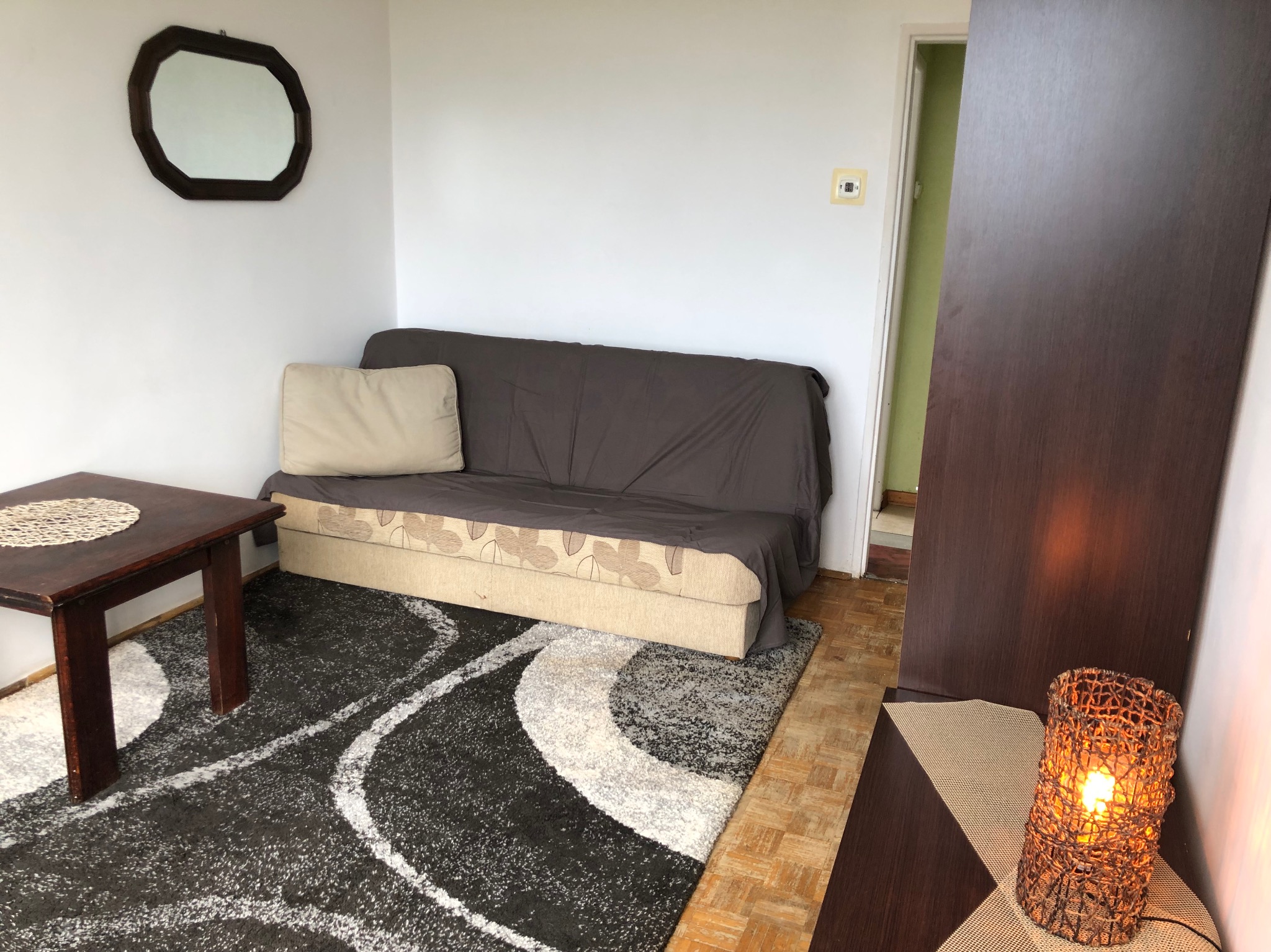Pokój dzienny z rozłożoną sofą przykrytą brązową narzutą, wzorzystym dywanem w czarno-białe fale, drewnianym stolikiem kawowym i lampą z plecionki, widoczne również lustro na ścianie i otwarte drzwi.
