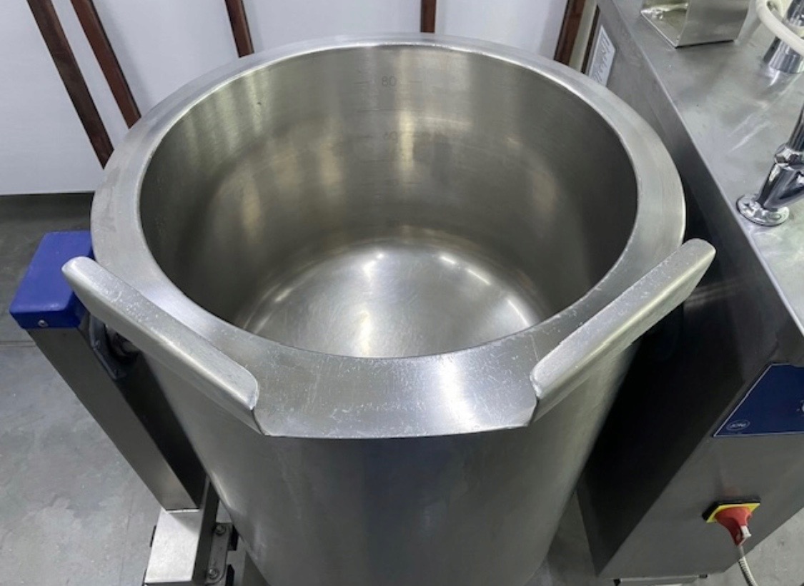 Wnętrze przemysłowego, nierdzewnego kotła warzelnego z widoczną miarką pojemności, umieszczonego na metalowej konstrukcji z elementami sterującymi.