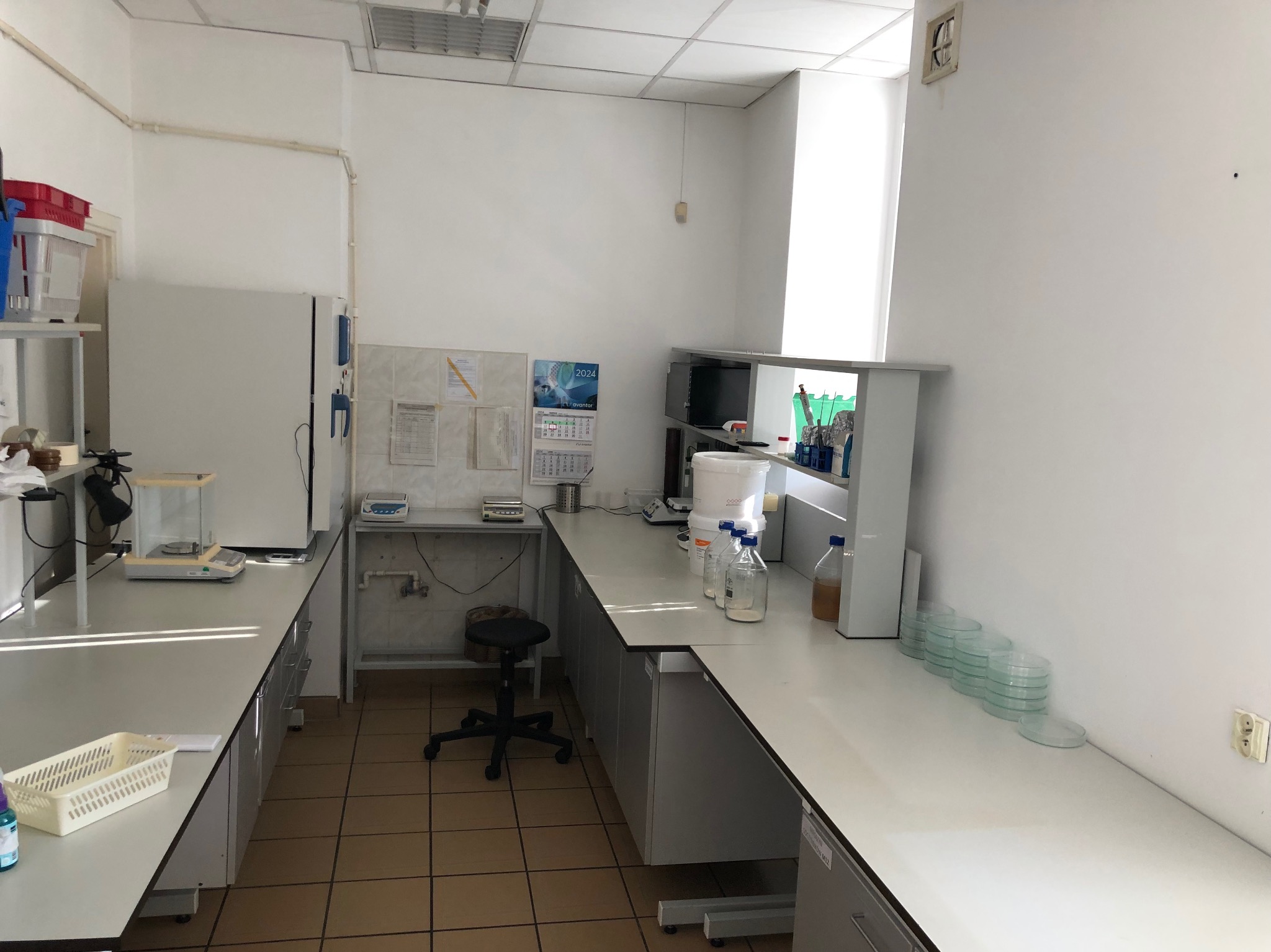 Jasne pomieszczenie laboratoryjne z białymi blatami, wyposażone w sprzęt, w tym wagi, probówki i szalki Petriego. Na ścianie kalendarz z datą 2024.