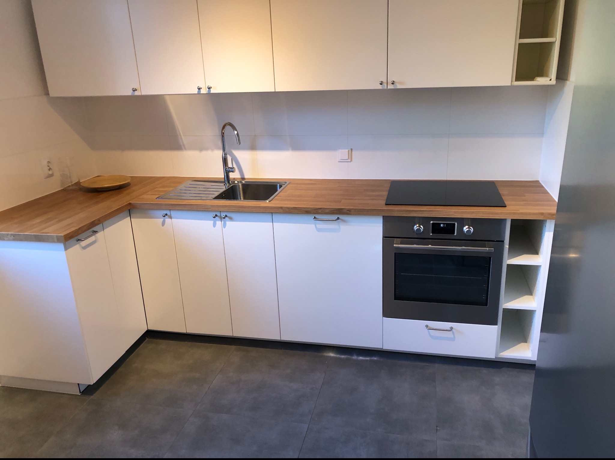 Nowoczesna, jasna kuchnia z białymi szafkami i drewnianym blatem, wyposażona w zintegrowany piekarnik i płytę indukcyjną, widoczna umywalka i bateria.