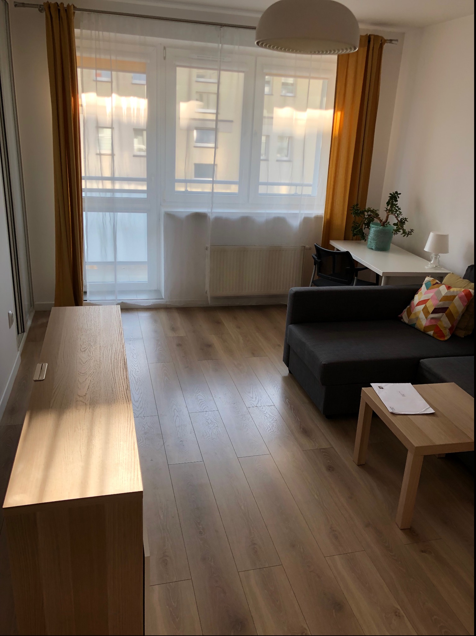 Jasne wnętrze pokoju dziennego z drewnianą podłogą, szarą sofą z kolorową poduszką, drewnianym stolikiem kawowym z dokumentami, biurkiem z rośliną i lampką oraz oknem z żółtymi zasłonami.