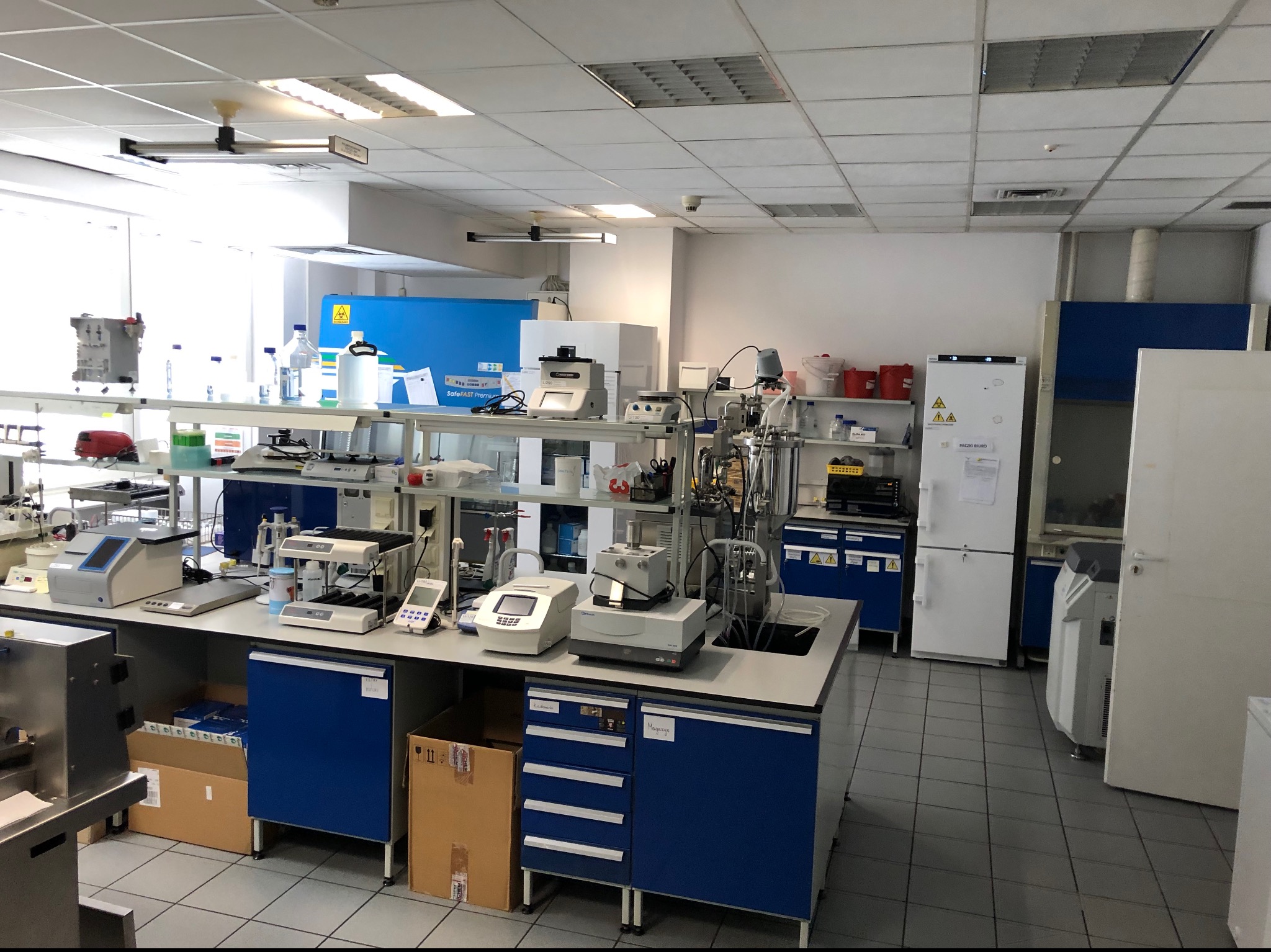 Szeroki kadr laboratorium z wyposażeniem: stoły laboratoryjne z urządzeniami pomiarowymi, szafki z szufladami, lodówka laboratoryjna i inne specjalistyczne sprzęty.