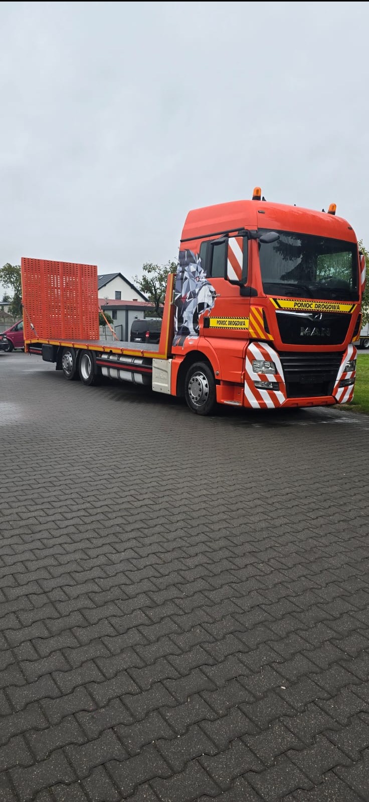 Pomarańczowa laweta MAN z grafiką na kabinie, zaparkowana na kostce brukowej.